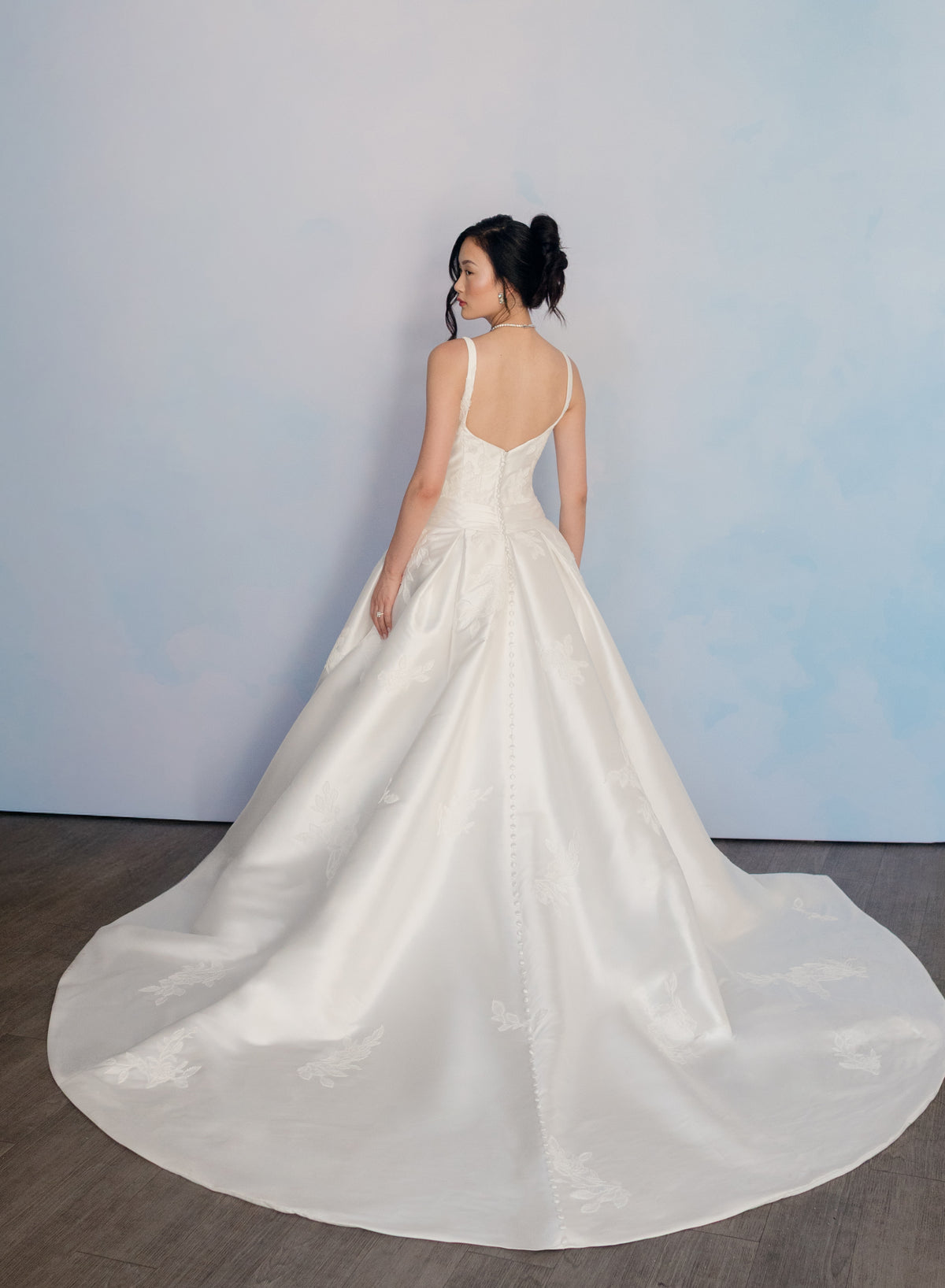 A woman wearing a white satin wedding gown with a backless open back and a full train, photographed from behind against a pale blue wall and dark wood floor. / Drop waist, mikado ballgown with pleating detail and scattered lace applique.