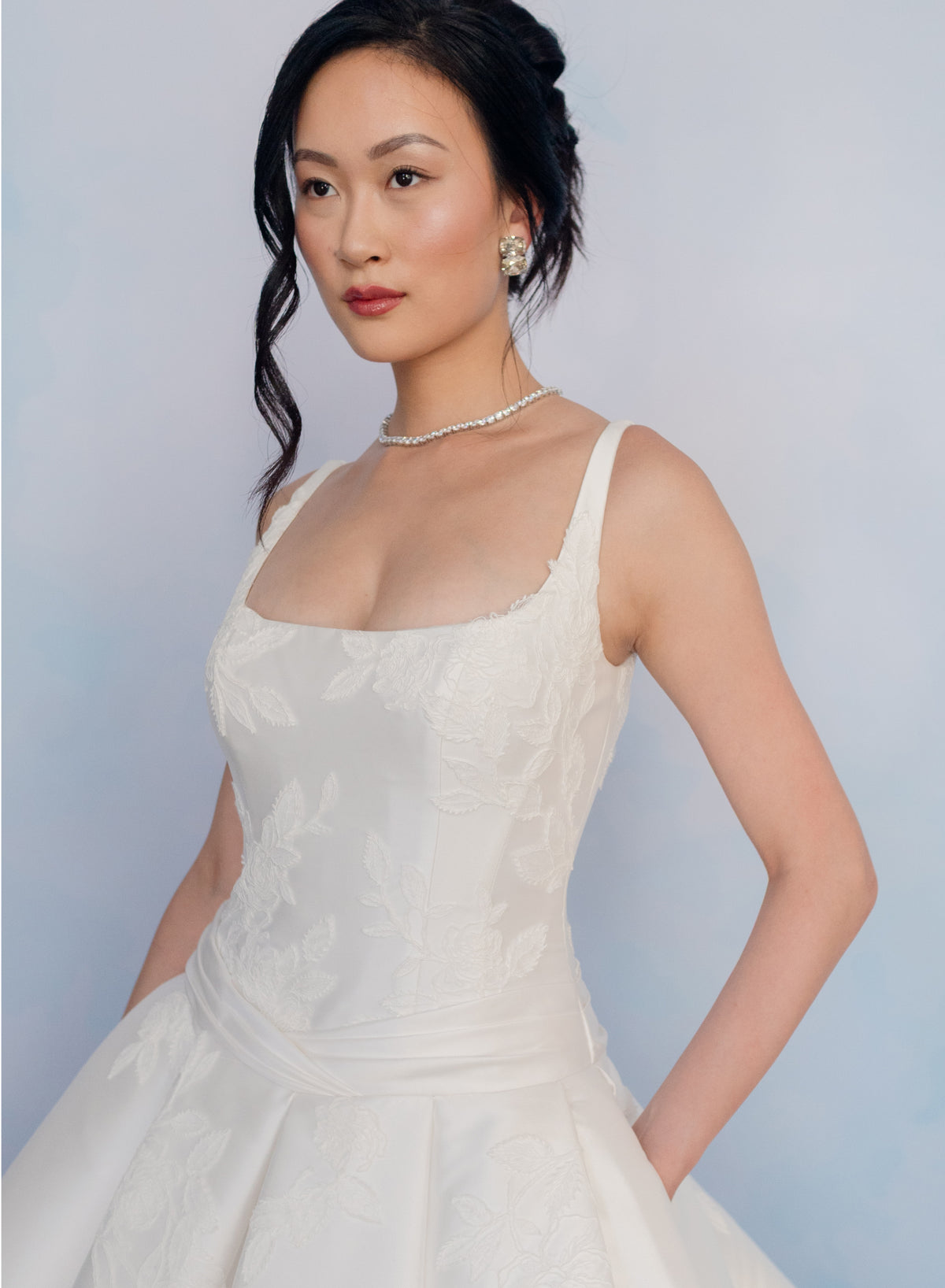 A woman wearing a white sleeveless lace dress with a square neckline and thin straps, posed against a soft light blue studio background. / Drop waist, mikado ballgown with pleating detail and scattered lace applique.
