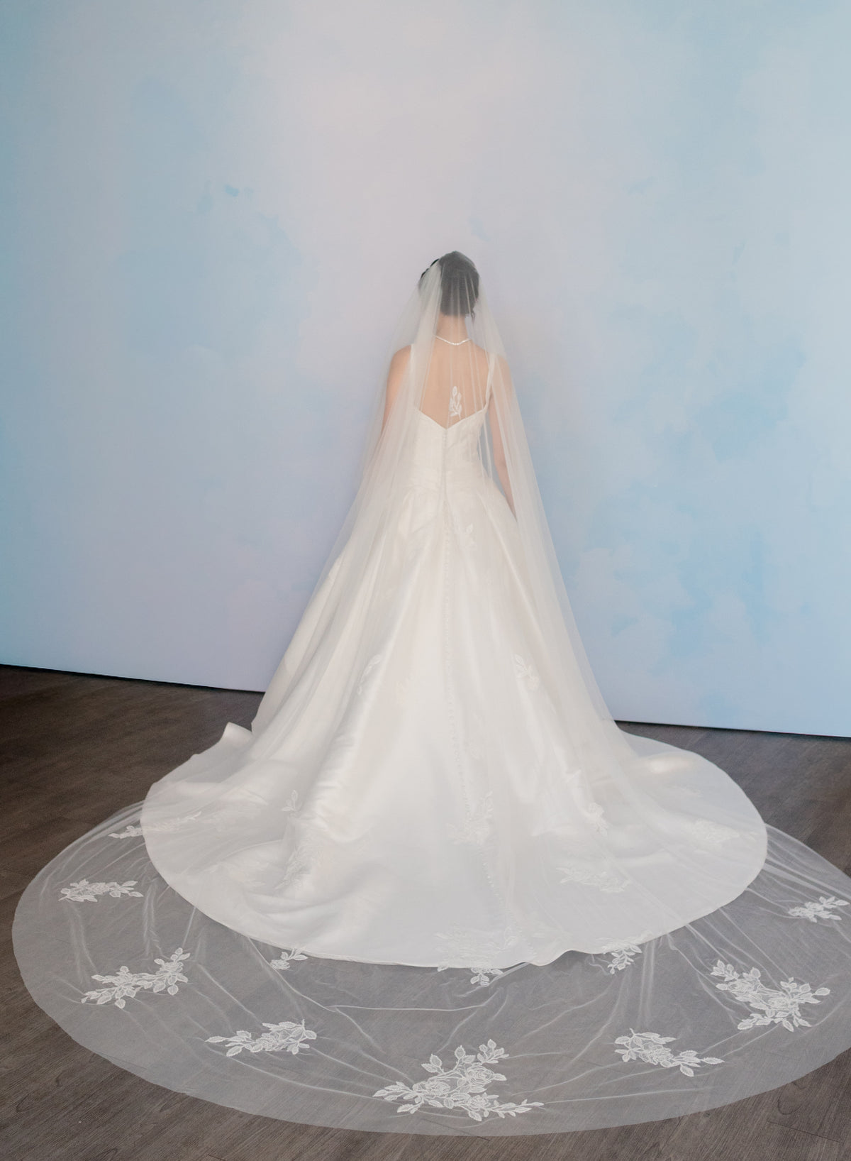 A bride viewed from behind wearing a white wedding dress with a long sheer veil featuring floral lace patterns, standing on a dark wood floor against a pale blue wall. / Lace and tulle veil.