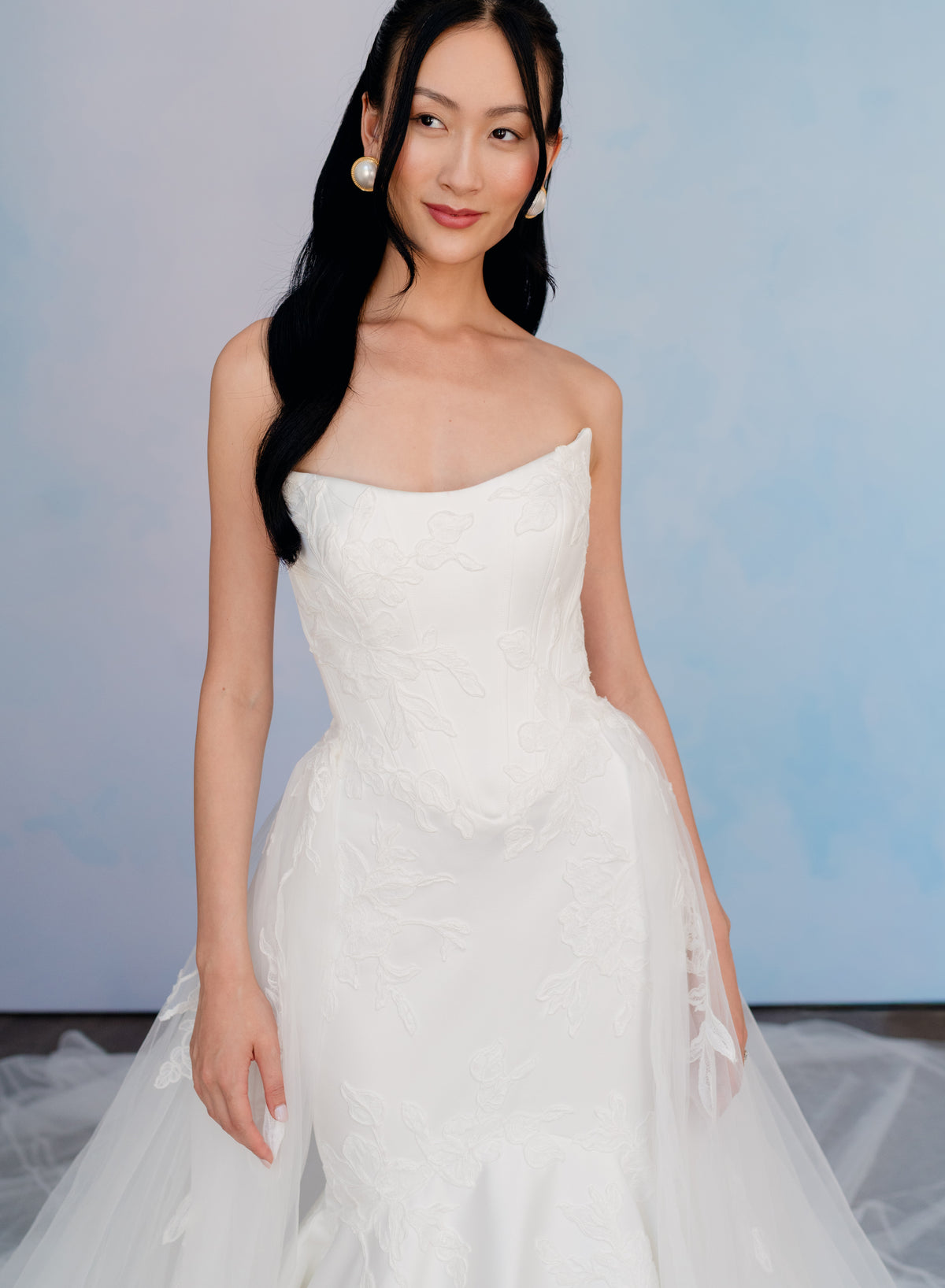 A woman wearing a strapless white wedding gown with a structured bodice and full tulle skirt, standing against a soft light blue studio backdrop. / Satin mermaid gown with lace appliques and detachable overskirt.