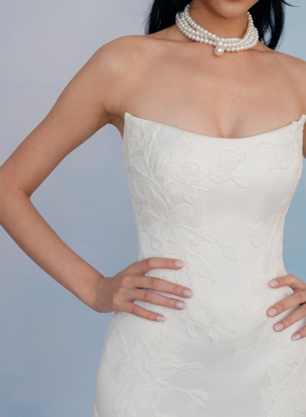 A close-up of a model wearing a strapless white lace dress with floral embroidery and a double-strand pearl necklace against a light blue background. / Satin mermaid gown with lace appliques and detachable overskirt.