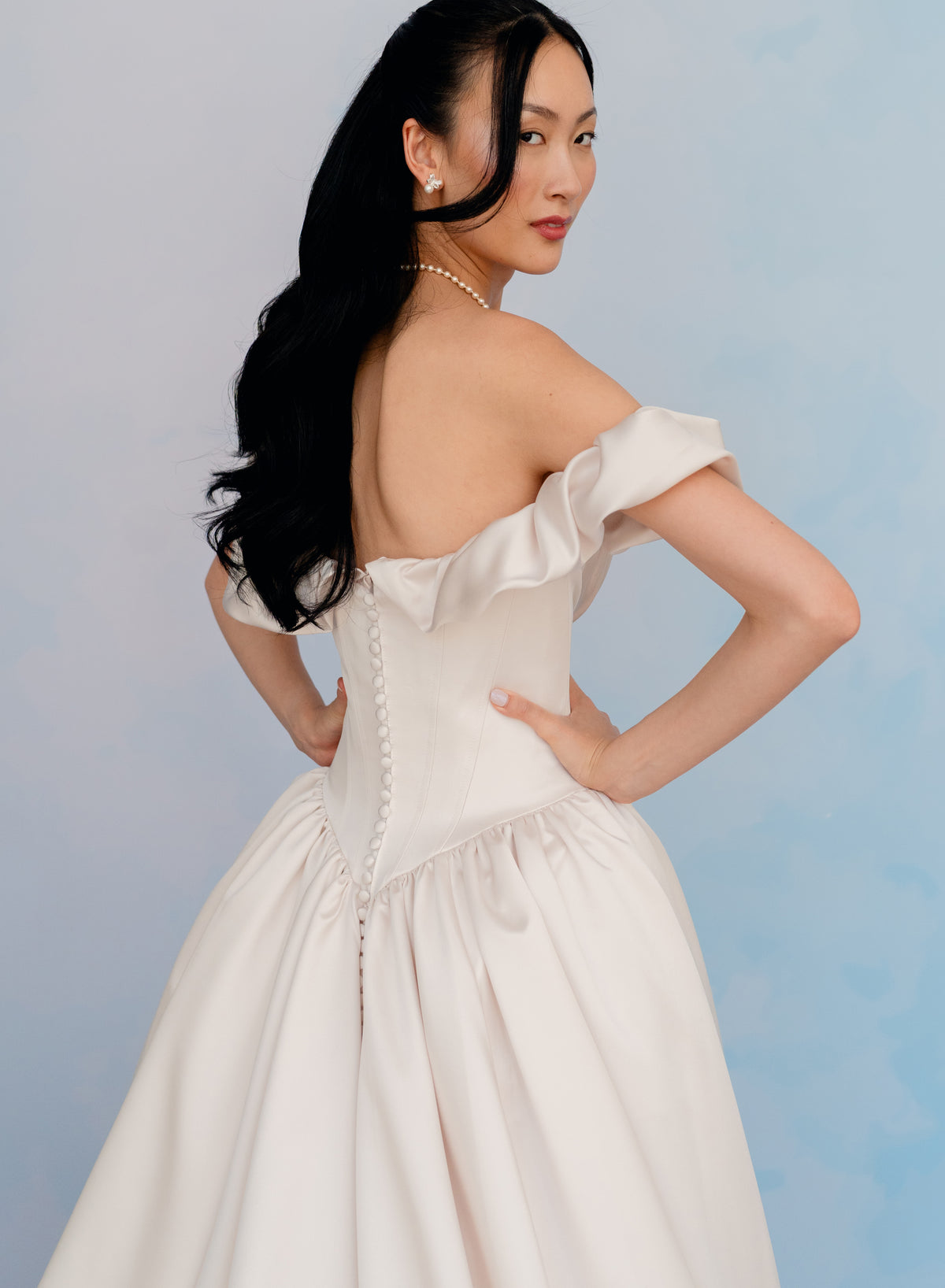 A woman modeling an off-the-shoulder cream satin gown with a structured corset-style bodice and full skirt, photographed against a soft light blue studio background. / Off the shoulder sculpted neckline gown with basque waist and full, gathered skirt.