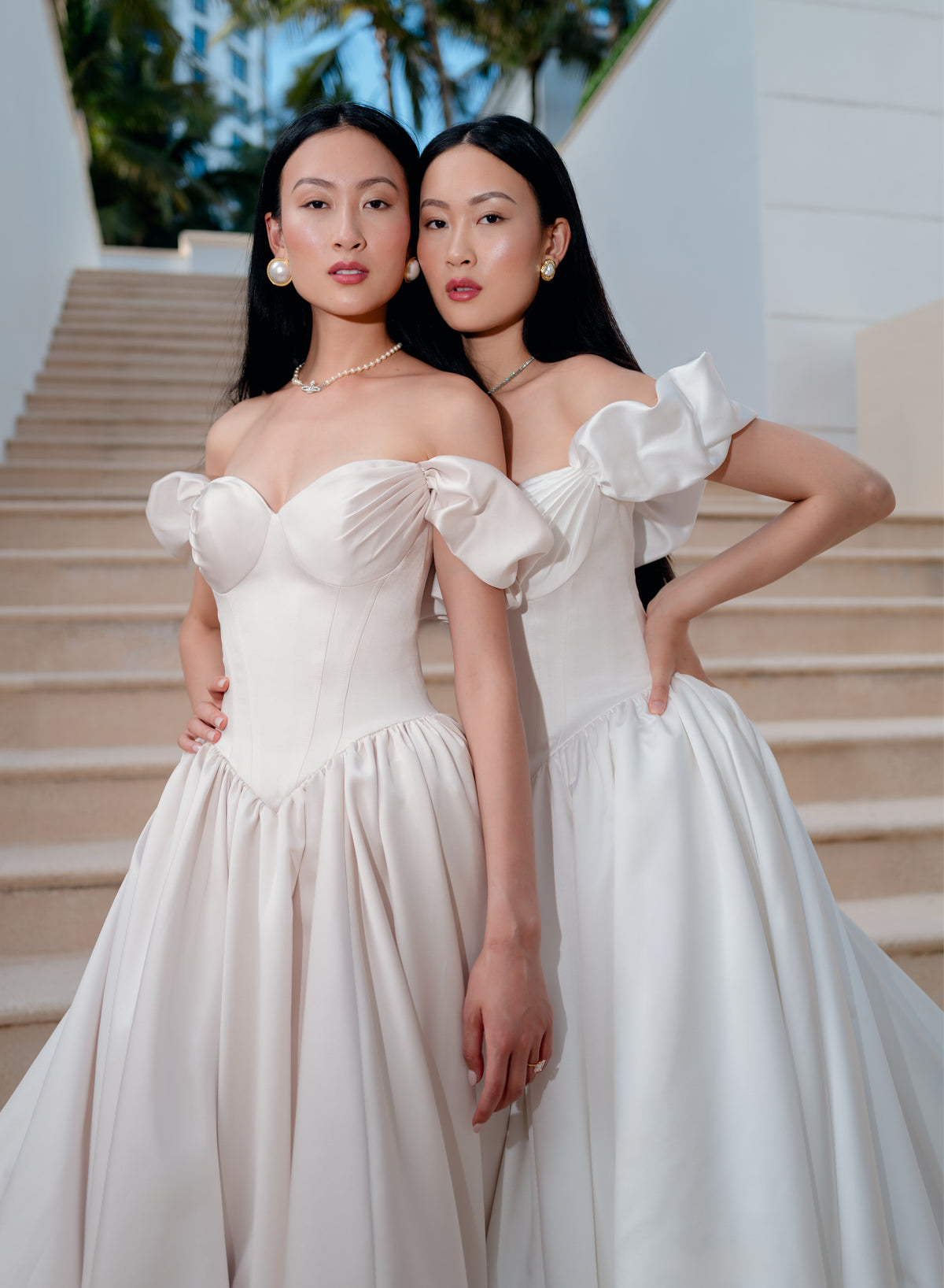 Two women wearing matching off-the-shoulder white ballgowns with structured puff sleeves and a fitted corset-style bodice, standing together on outdoor stone stairs. / Off the shoulder sculpted neckline gown with basque waist and full, gathered skirt.