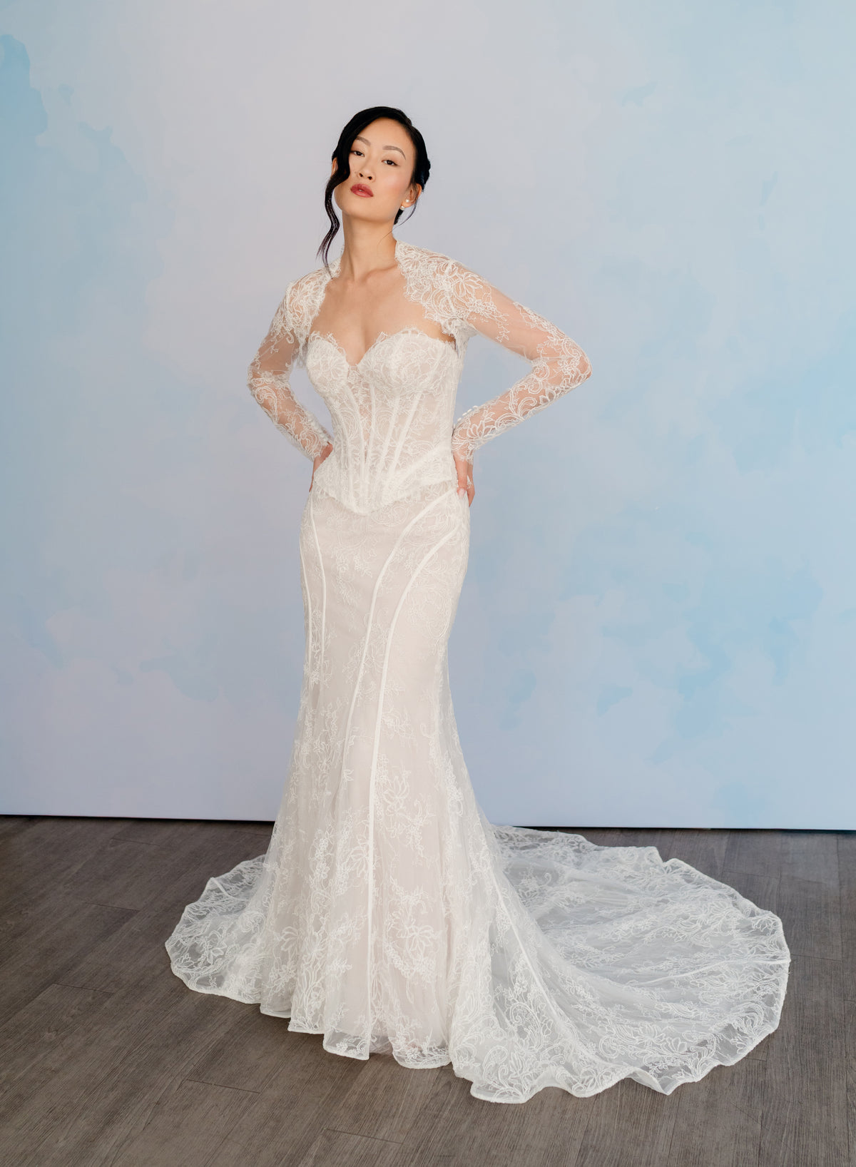 A woman modeling a fitted lace wedding gown with long sheer sleeves, a sweetheart neckline, and a train, standing against a pale blue studio backdrop. / Slim lace gown with semi-sheer bodice with sweetheart neckline and bustier cut. Small peplums on sides hips. Invisible zipper back closure. Matching lace bolero with long sleeves.