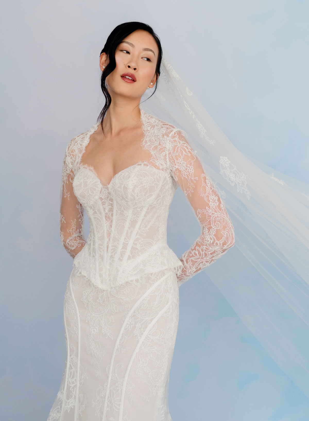 A woman modeling a white lace wedding dress with long sheer lace sleeves and a matching veil against a soft light blue studio background. / Long tulle veil with lace motifs scattered on top area.