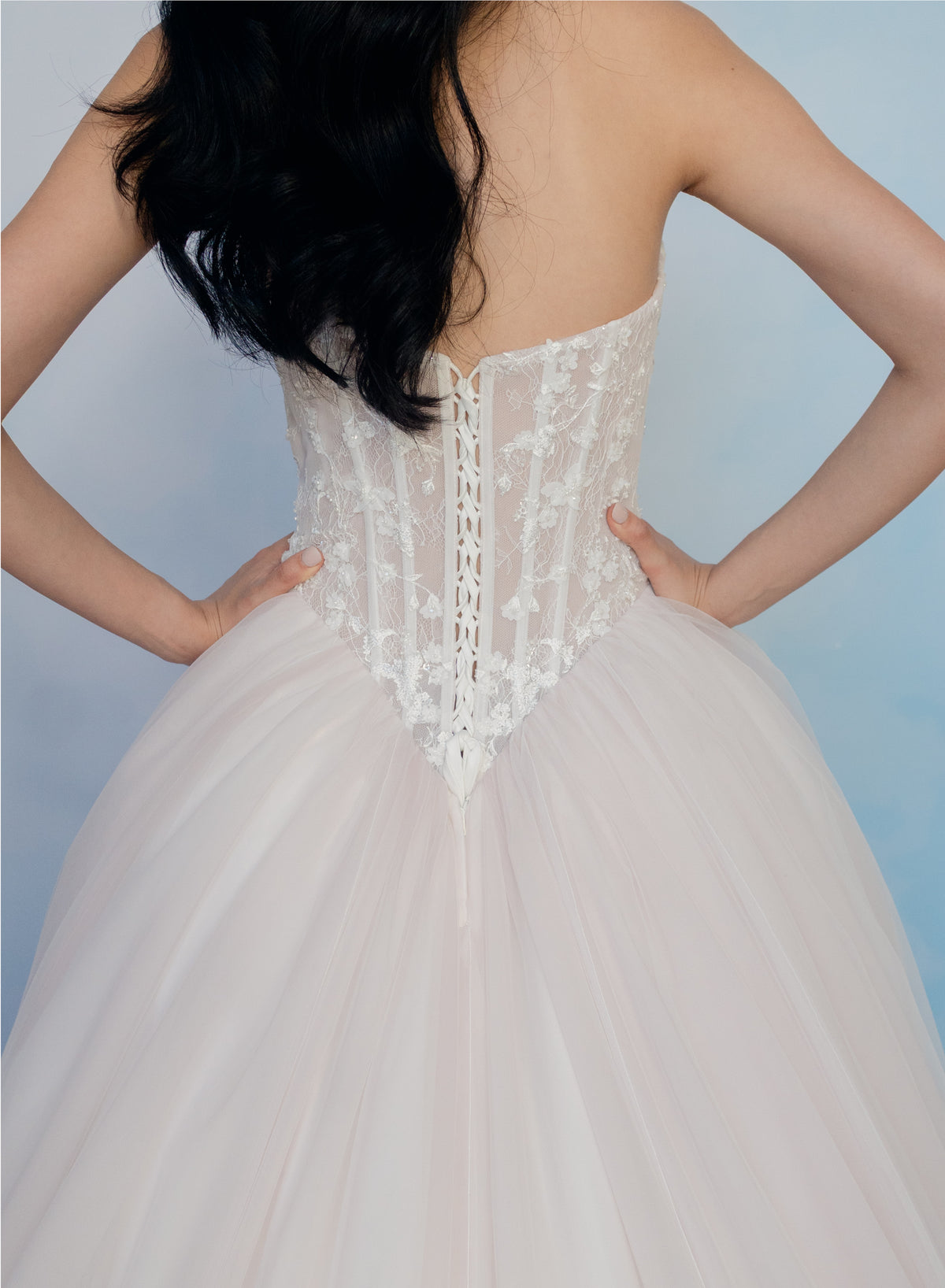 Back view of a strapless white wedding gown with a lace corset-style back and a full tulle skirt, shown on a model against a soft blue gradient background. / Semi sheer Basque waist bodice with lace appliques and dramatic tulle skirt.