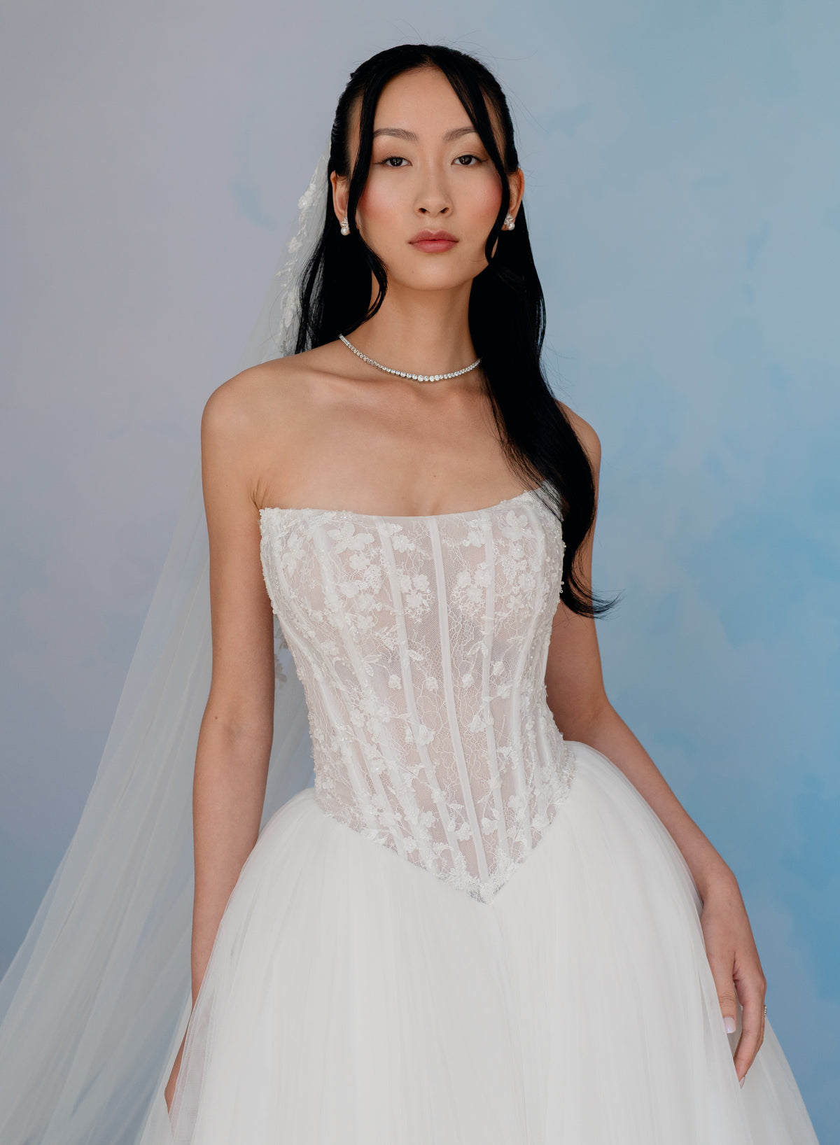 A woman modeling a strapless white lace wedding gown with a fitted lace bodice and a full tulle skirt, posed against a soft blue studio backdrop. / Semi sheer Basque waist bodice with lace appliques and dramatic tulle skirt.