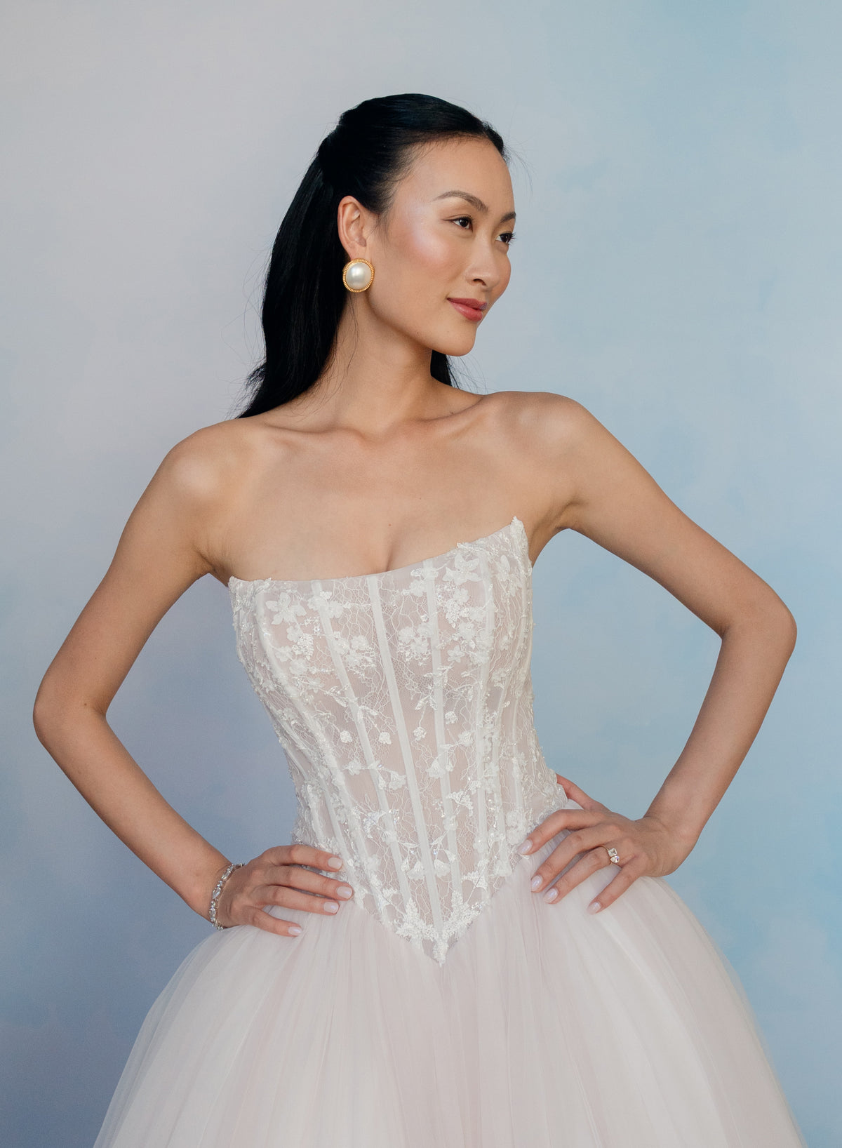 A woman modeling a strapless white lace corset dress with a voluminous tulle skirt, posed against a soft light blue studio backdrop. / Semi sheer Basque waist bodice with lace appliques and dramatic tulle skirt.