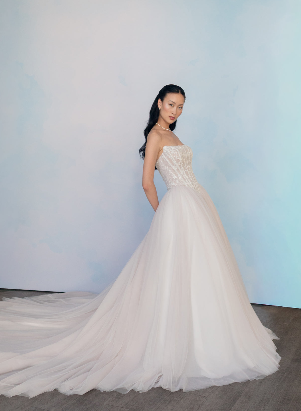 A woman modeling a strapless white wedding gown with a fitted embellished bodice and a voluminous tulle skirt with a long train, standing against a soft light blue studio backdrop. / Semi sheer Basque waist bodice with lace appliques and dramatic tulle skirt.
