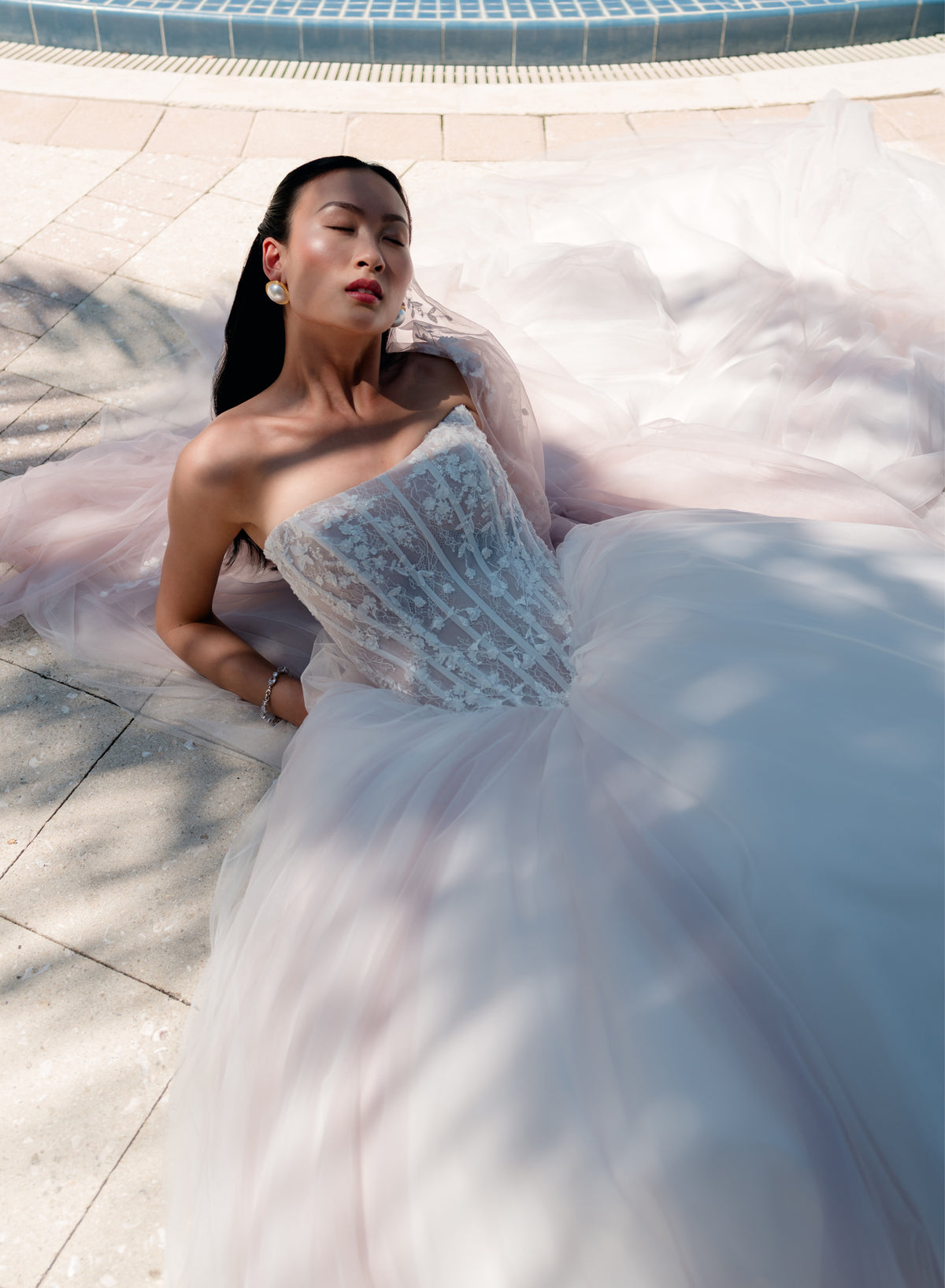 A woman reclines outdoors beside a tiled pool wearing a voluminous strapless tulle gown with a heavily embellished lace corset bodice. The dress is pale blush pink and white, with dramatic sunlight and shadows across the fabric. / Semi sheer basque waist bodice with lace appliques and dramatic tulle skirt.