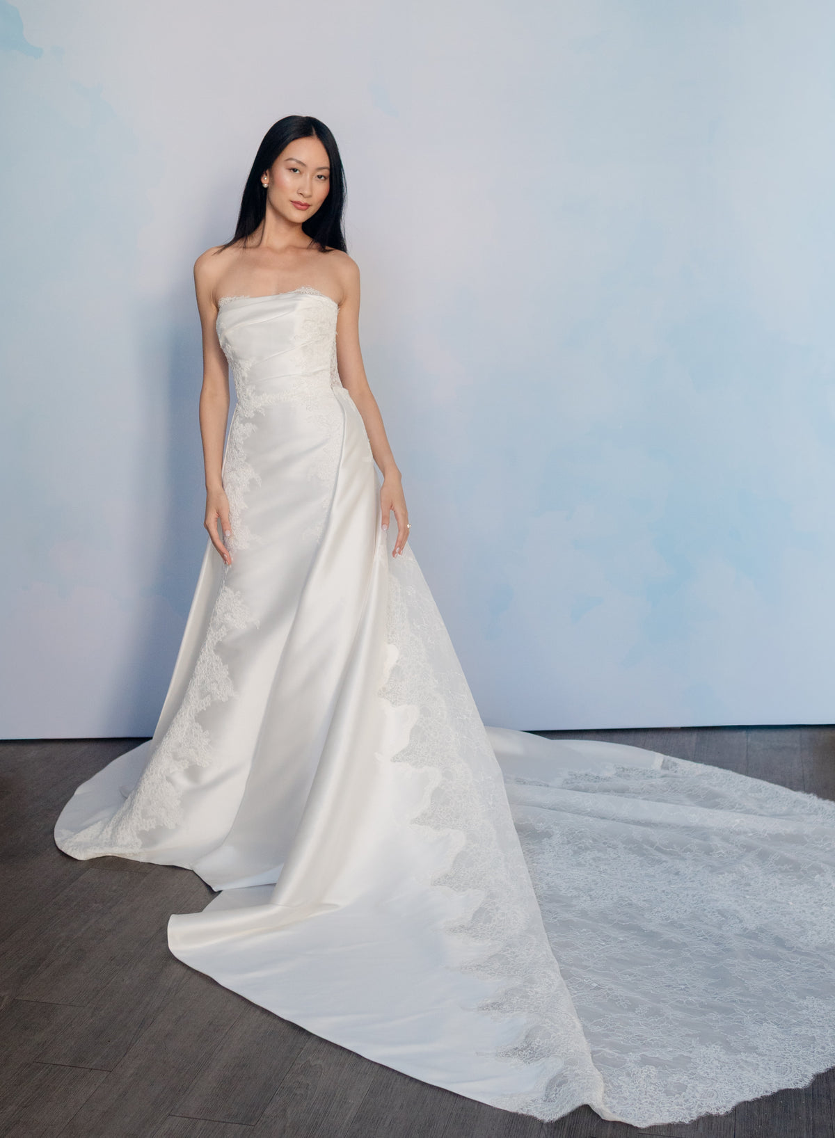 A woman modeling a strapless white satin wedding gown with lace applique and a long train against a soft blue studio backdrop. / Slim mikado style with pleated bodice with straight neckline and zipper/buttons back closure. Lace scallop decoration on bodice and on skirt sides. Matching mikado detachable train with sheer back area covered with lace.
