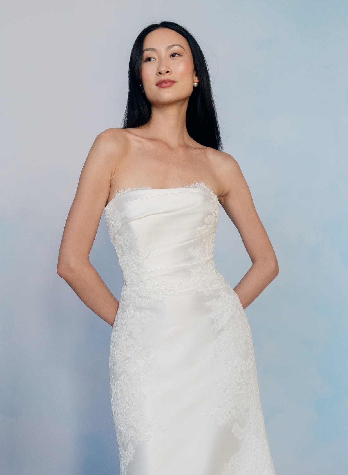 A woman modeling a strapless white wedding gown with lace applique and a fitted silhouette, standing against a soft light blue studio background. / Slim mikado style with pleated bodice with straight neckline and zipper/buttons back closure. Lace scallop decoration on bodice and on skirt sides. Matching mikado detachable train with sheer back area covered with lace.