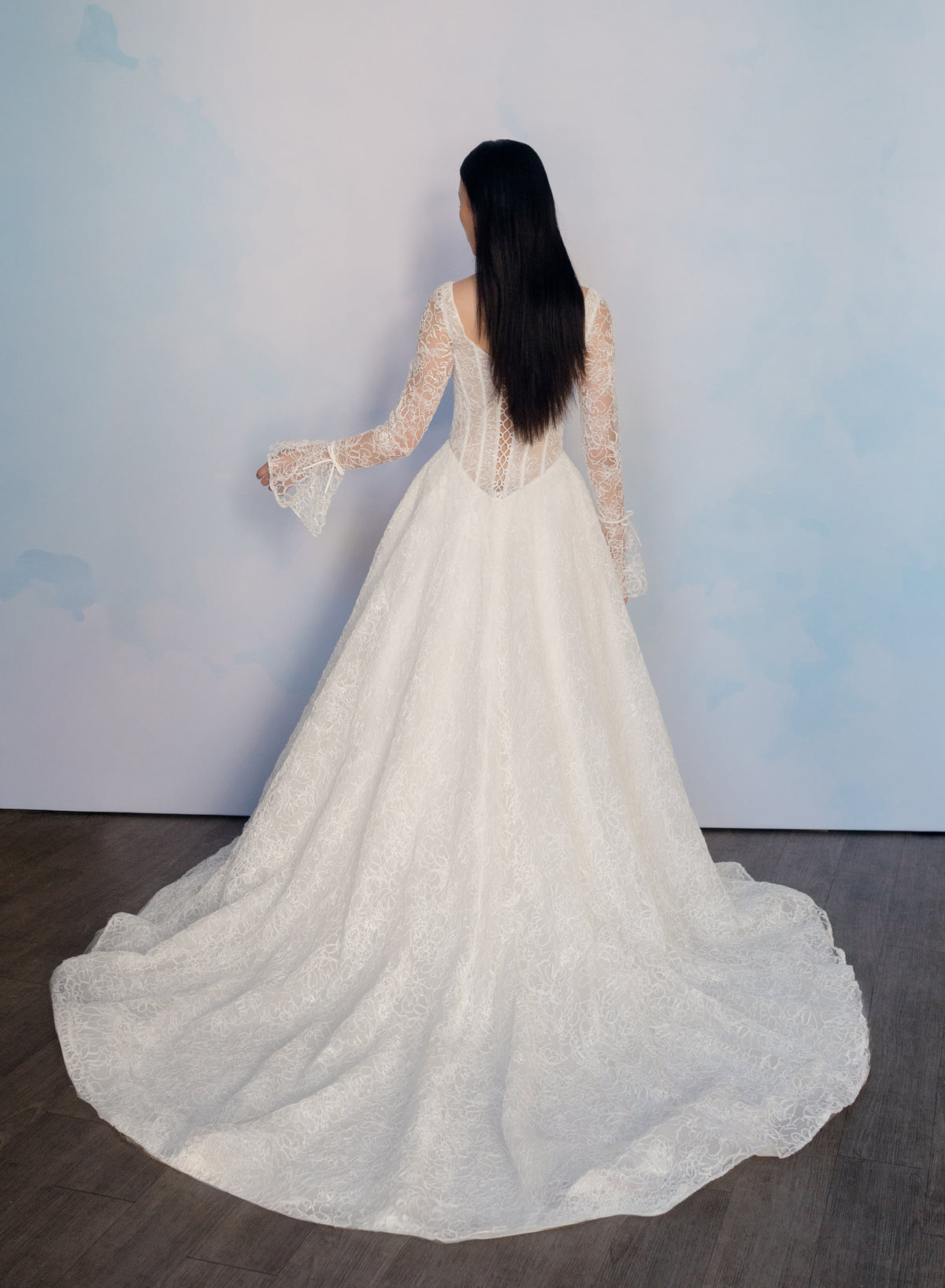 A woman viewed from behind wearing a white lace wedding gown with long sheer sleeves and a full train, standing against a pale blue wall on a dark wood floor. / All over lace gown with long sleeve, basque waist and A-line skirt with lace up back.