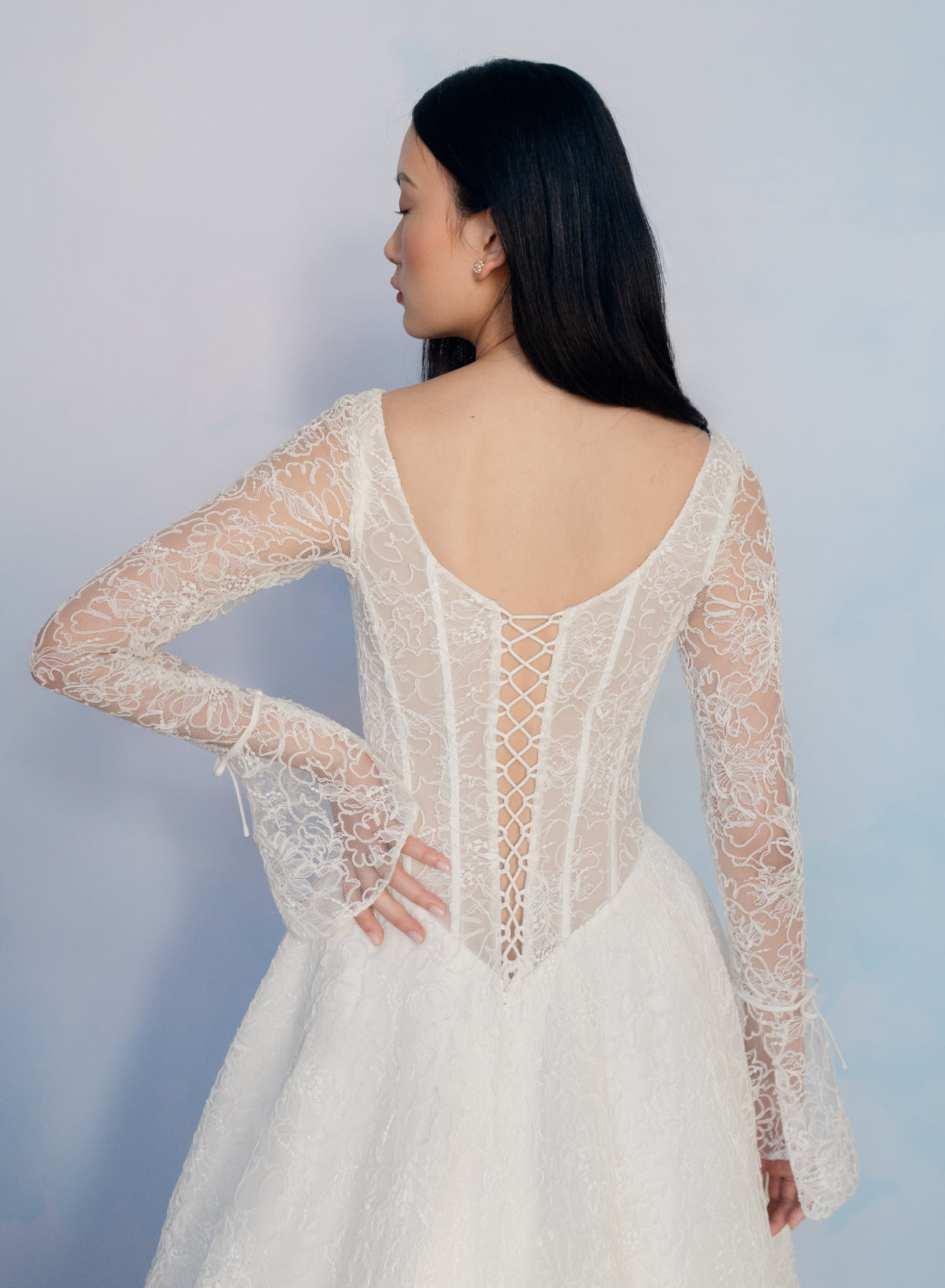 A woman wearing a white lace bridal gown, shown from the back. The dress features long sheer lace sleeves, a deep V-back, and an open lace-up corset-style back. / All over lace gown with long sleeve, basque waist and A-line skirt with lace up back.