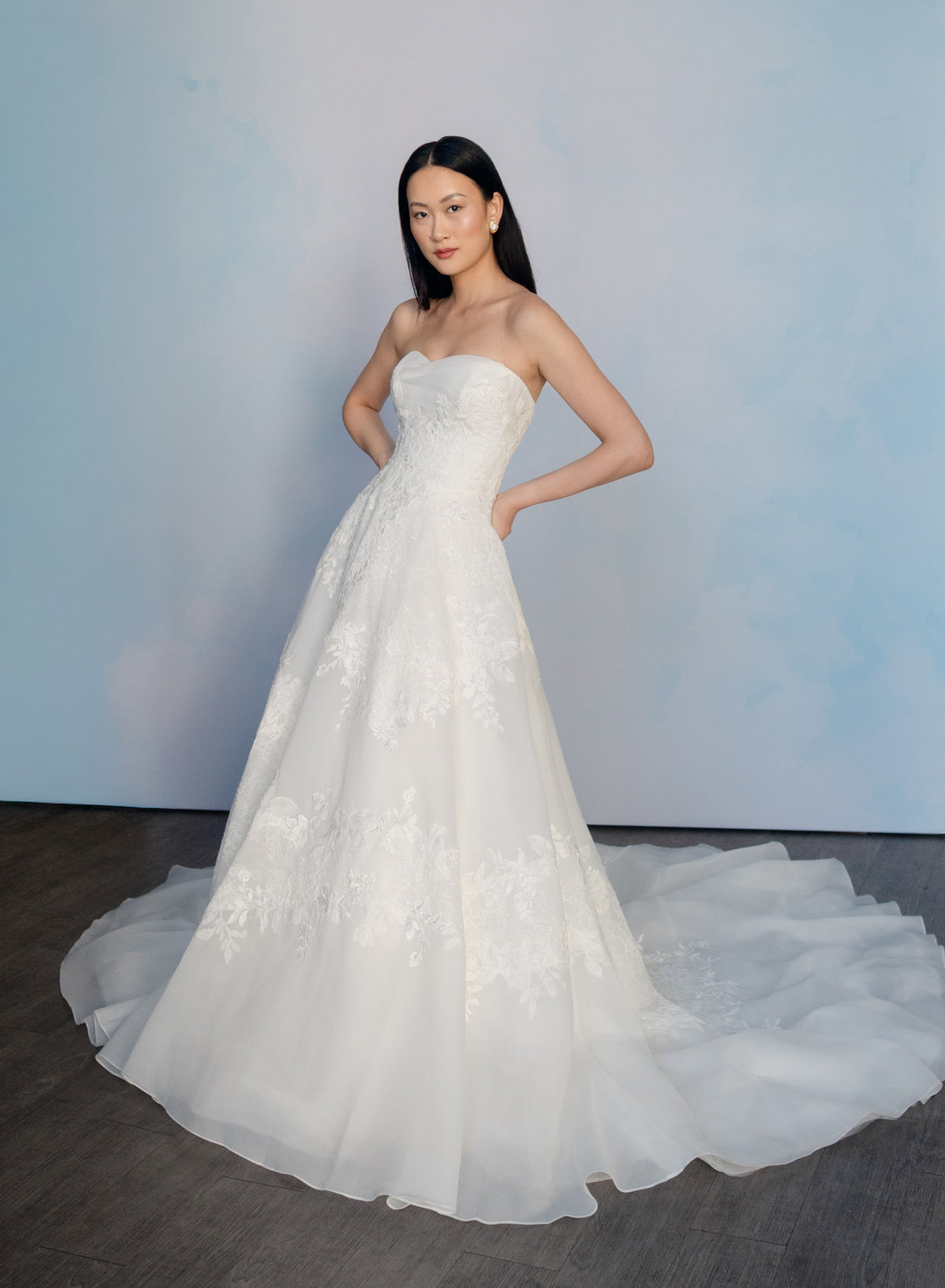A woman modeling a strapless white A-line wedding gown with floral lace applique and a full tulle train, standing against a soft blue studio backdrop. / Organza and lace princess ballgown with lace applique details.