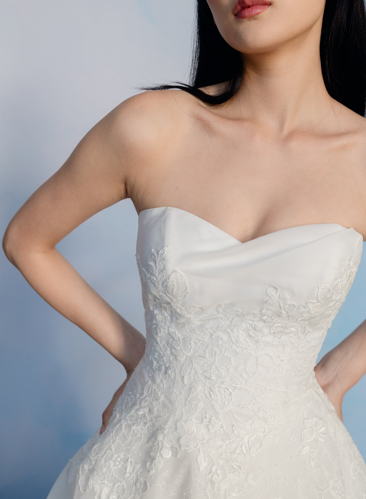 A close-up of a model wearing a strapless white wedding dress with a sweetheart neckline, smooth satin-like fabric, and floral lace applique on the bodice, against a soft blue studio background. / Organza and lace princess ballgown with lace applique details.
