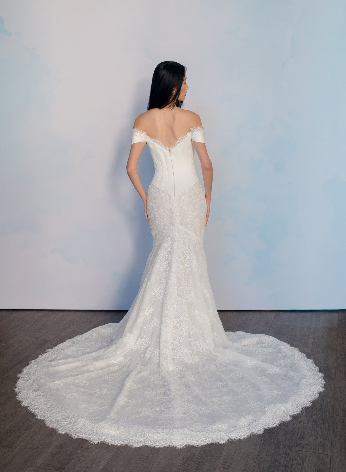 A woman stands with her back to the camera wearing an off-the-shoulder lace wedding gown with a long train, set against a light blue textured wall and dark wood floor. / Slim style mock 2 piece with satin bodice and semi-sheer lace skirt. Scooped neckline with off-shoulder straps decorated with delicate lace scallop edge, invisible zipper back closure.