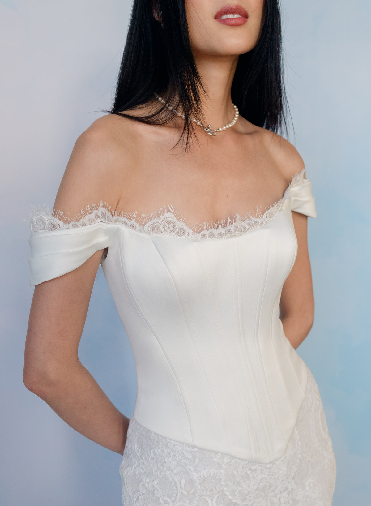 An off-the-shoulder white satin corset top with delicate lace trim along the neckline, worn by a model against a soft blue gradient background. / Slim style mock 2 piece with satin bodice and semi-sheer lace skirt. Scooped neckline with off-shoulder straps decorated with delicate lace scallop edge, invisible zipper back closure.