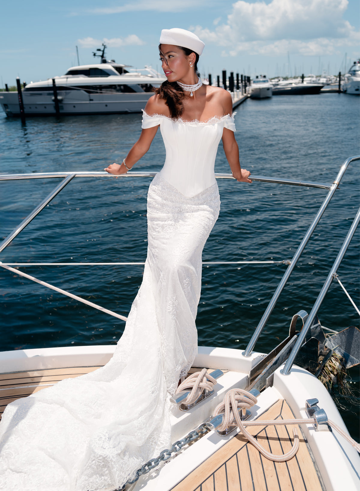 A woman wearing a white off-the-shoulder wedding gown and matching white sailor hat stands on the bow of a yacht, with water, docks, and other boats in the background. / Slim style mock 2 piece with satin bodice and semi-sheer lace skirt. Scooped neckline with off-shoulder straps decorated with delicate lace scallop edge, invisible zipper back closure.