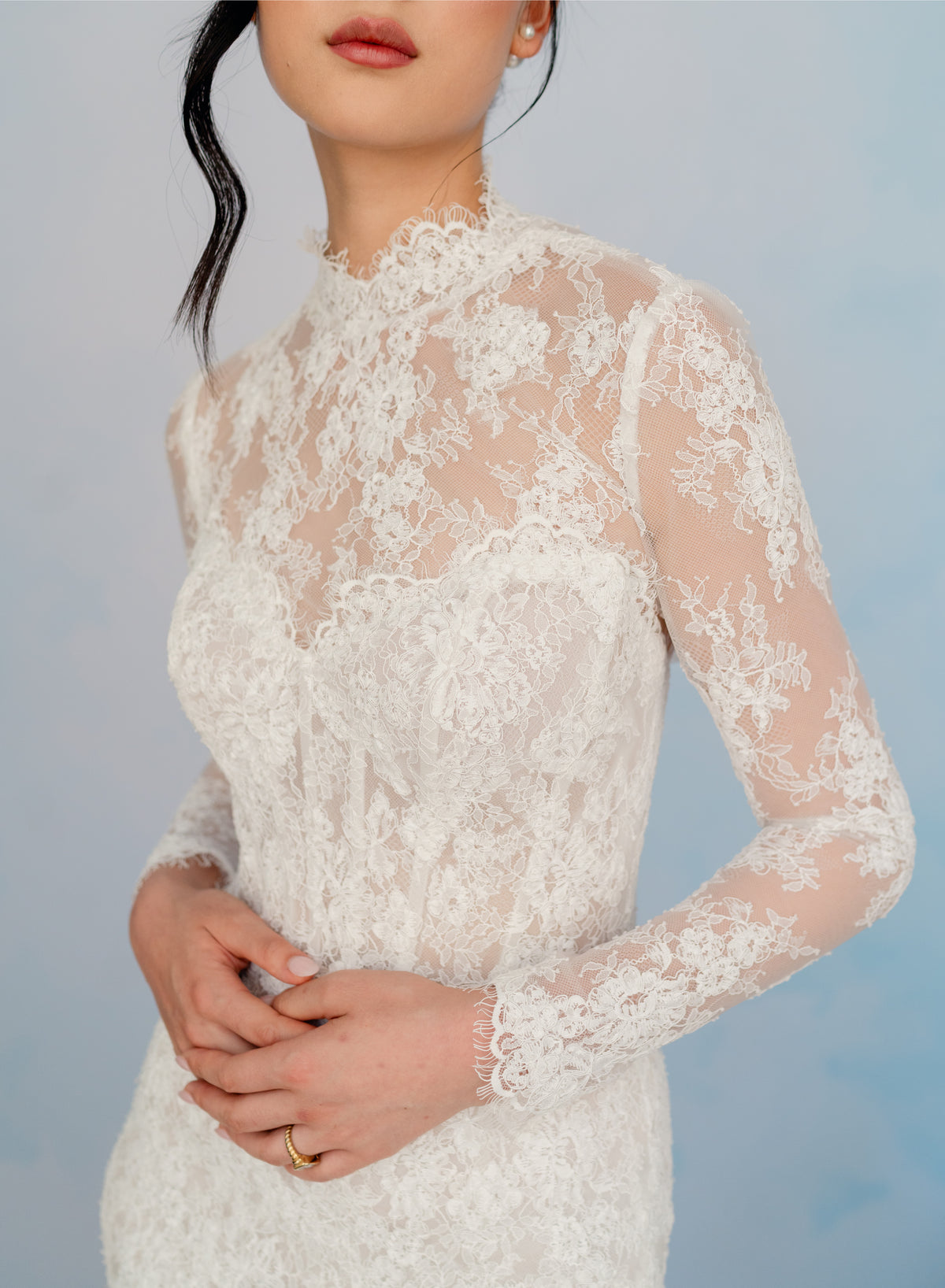 A woman wearing a white lace long-sleeve wedding dress with a sheer mesh bodice and floral lace sleeves, shown against a soft light blue studio background. / All over lace and lace applique slim gown with detachable lining and jacket as well as structured corset.