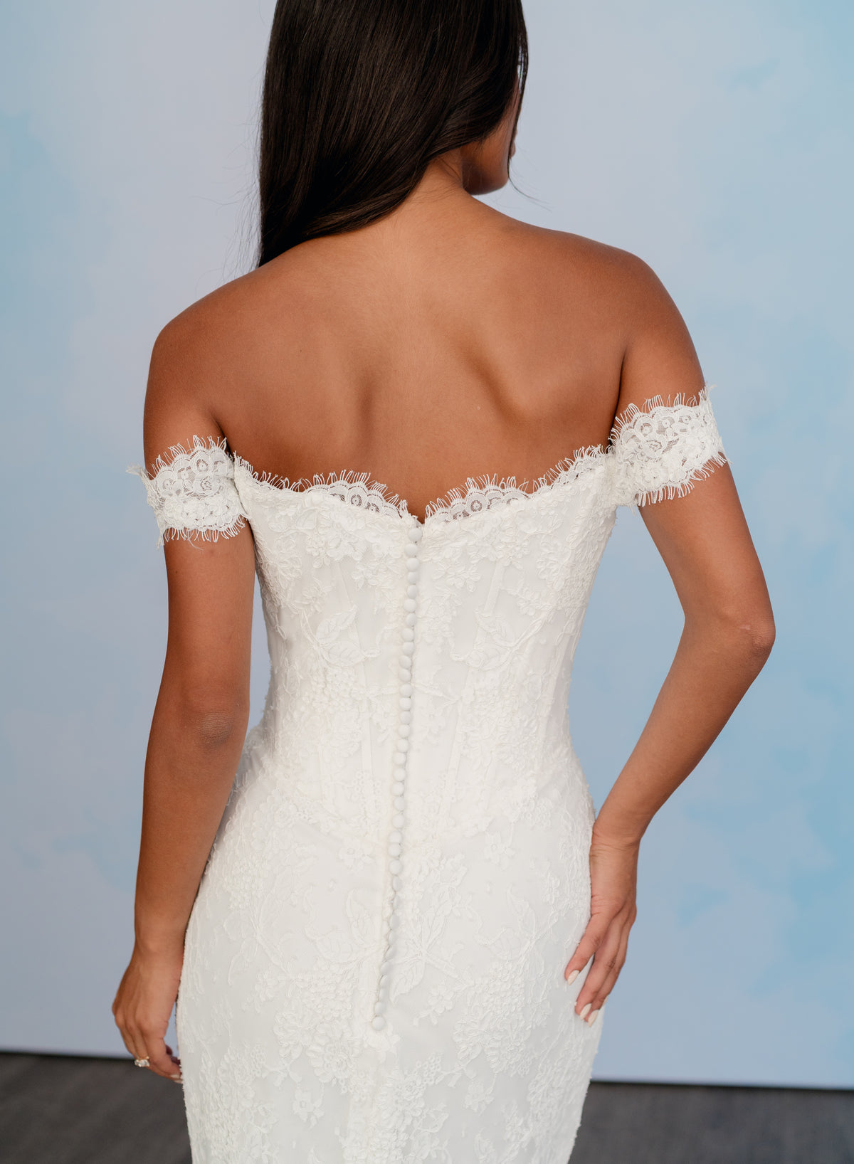 A woman shown from the back wearing an off-the-shoulder lace wedding dress with a fitted bodice and lace trim at the neckline and sleeves, against a light blue studio background. / All over lace column gown with a dramatic matching overskirt.