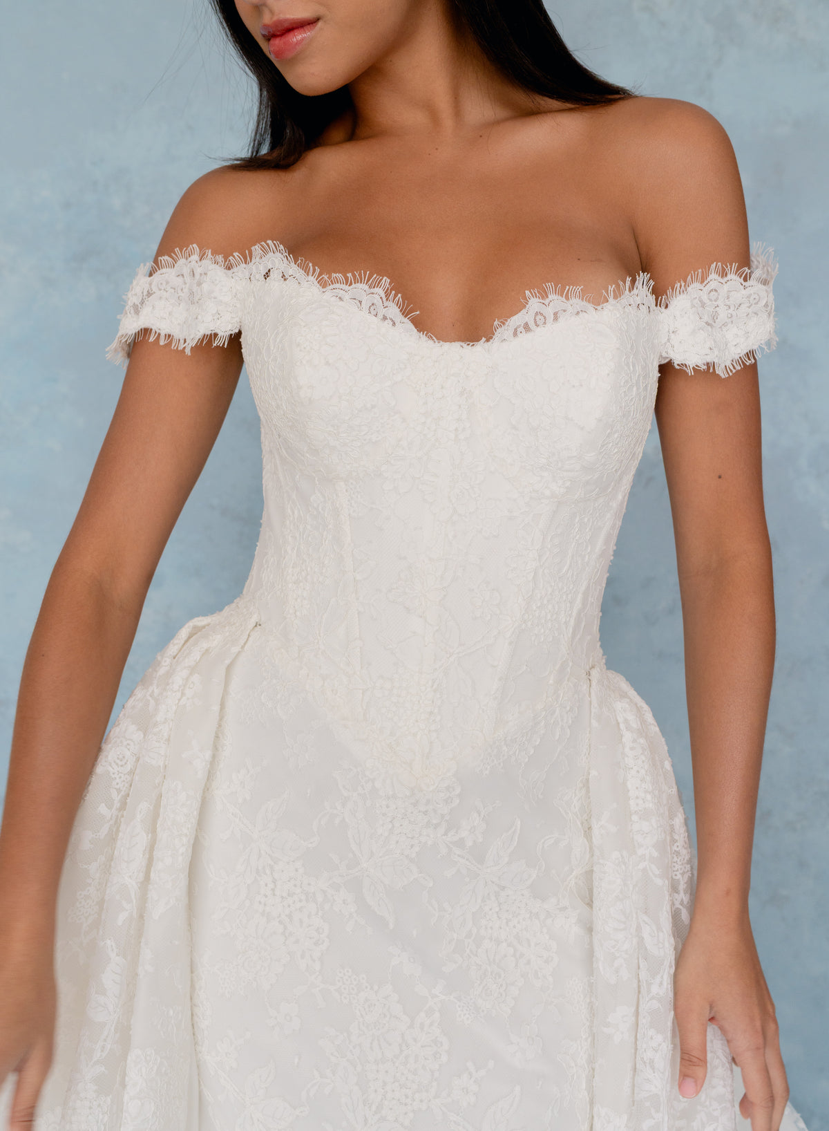 An off-the-shoulder white lace dress with a fitted corset-style bodice and a full skirt, shown on a model against a textured light blue backdrop. / All over lace column gown with a dramatic matching overskirt.