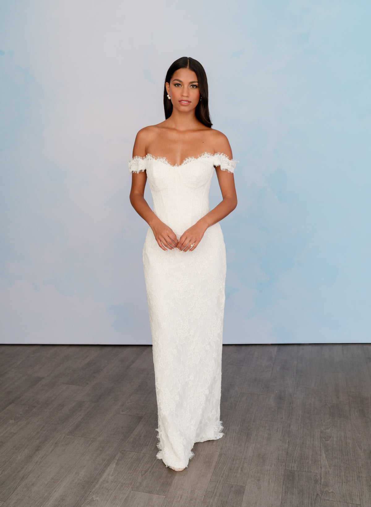 A woman modeling an off-the-shoulder white lace wedding gown with a fitted bodice and floor-length skirt, standing against a light blue studio backdrop with a gray wood floor. / All over lace column gown with a dramatic matching overskirt.