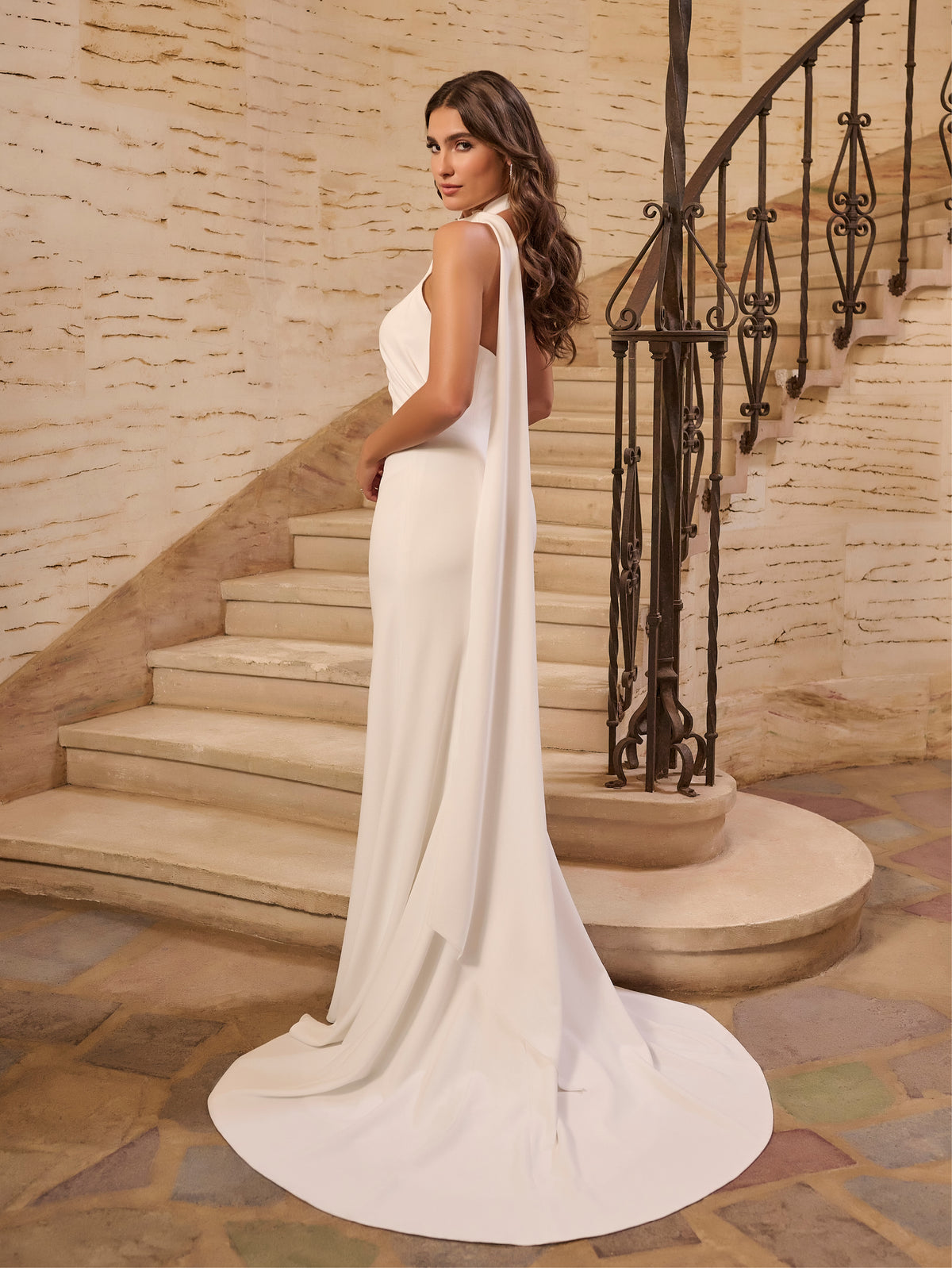 Woman in a white gown standing on a staircase with stone walls. / Halter stretch crepe trumpet with self scarf.