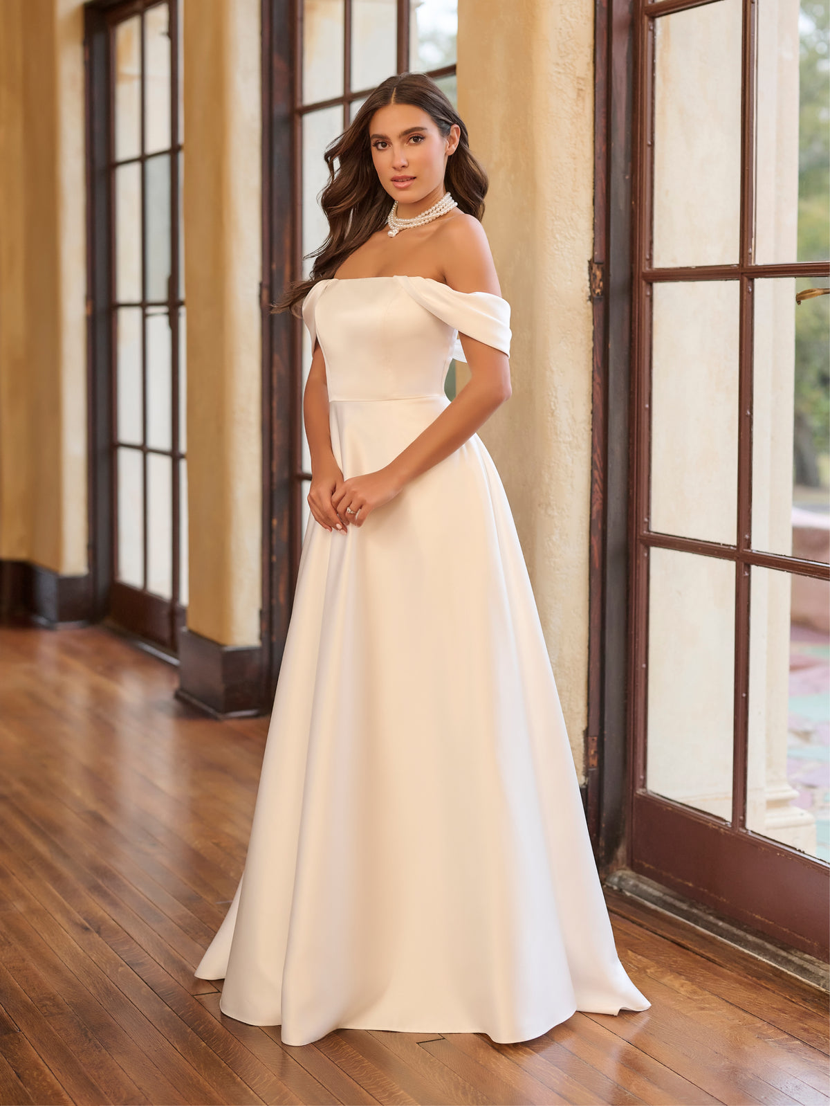 Woman in a white off-shoulder gown standing in a room with large windows. / Draped off the shoulder satin A-line.