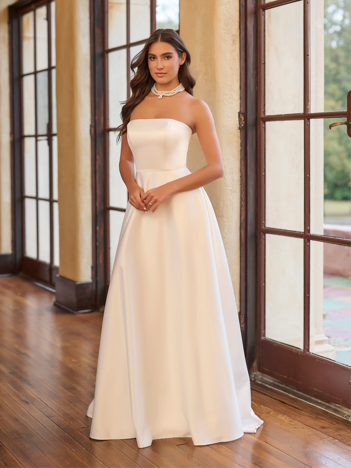 Woman in a white strapless gown standing indoors with large windows in the background / Draped off the shoulder satin A-line.