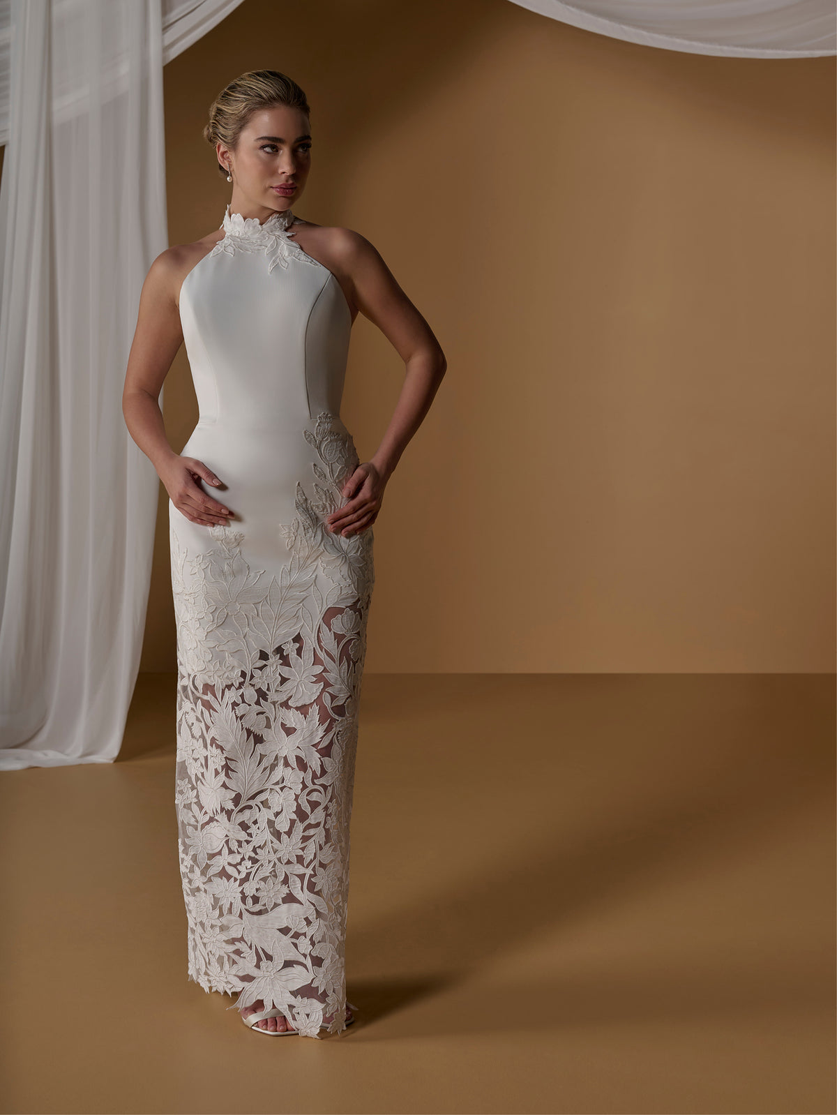 A woman models a sleeveless, high-neck floor-length wedding gown with a fitted bodice and intricate floral lace overlay on the skirt, posed against a warm tan studio backdrop with draped white fabric. / Unlined skirt. Column gown with halter neckline and lace detailing.
