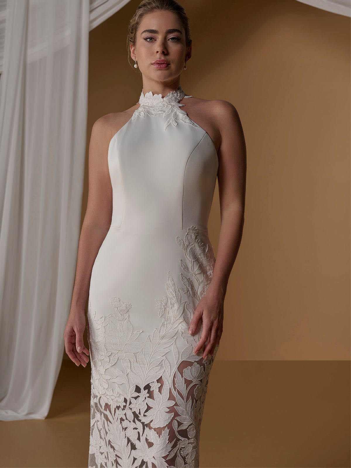 A woman modeling a sleeveless halter-neck white dress with floral lace applique at the high neckline and along the lower skirt, posed against a warm tan studio backdrop with draped white fabric on the left. / Unlined skirt. Column gown with halter neckline and lace detailing.