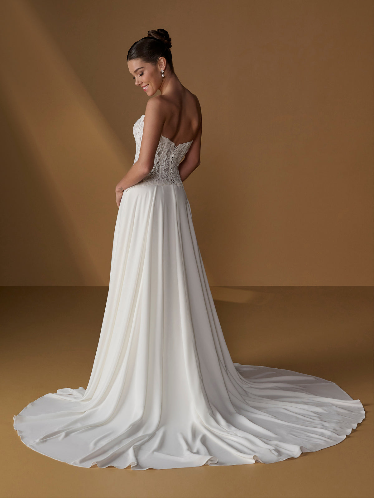 A woman modeling a strapless white wedding gown with a fitted lace bodice and a long flowing train, photographed in a warm brown studio setting. / Faux 2-piece gown with lace bodice and full skirt.
