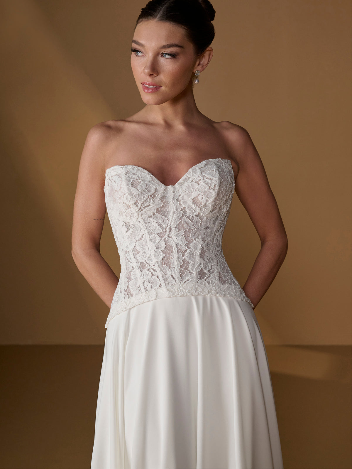 A woman modeling a strapless wedding gown with a lace corset-style bodice and a flowing ivory skirt, posed against a warm tan studio backdrop. / Faux 2-piece gown with lace bodice and full skirt.