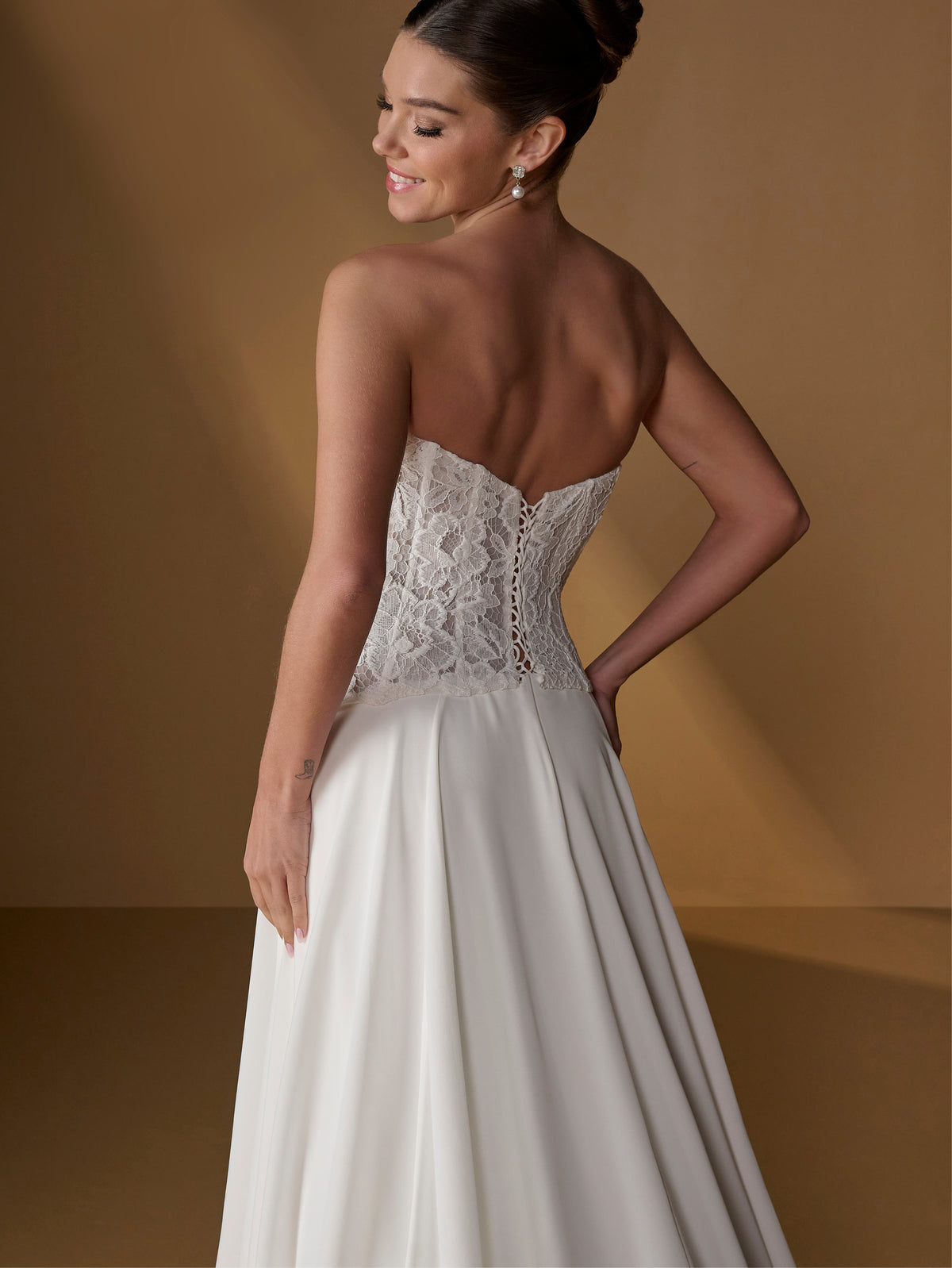 A woman modeling a strapless white wedding gown from the back, showing a lace corset-style bodice and flowing satin skirt against a warm brown studio backdrop. / Faux 2-piece gown with lace bodice and full skirt.