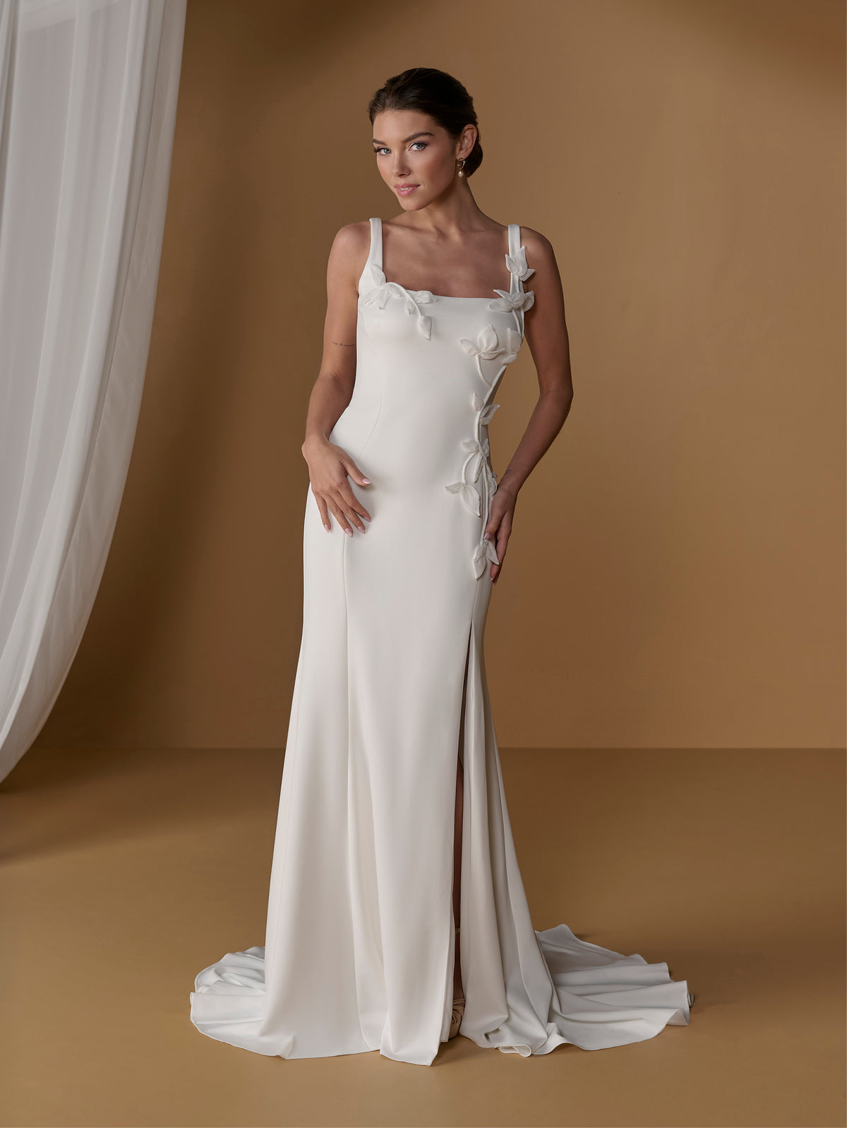 A woman modeling a floor-length ivory wedding gown with thin straps, a square neckline, side bows, and a slight train, posed against a warm tan studio backdrop with draped fabric at the left. / Square neckline scuba gown with handmade chiffon flowers.