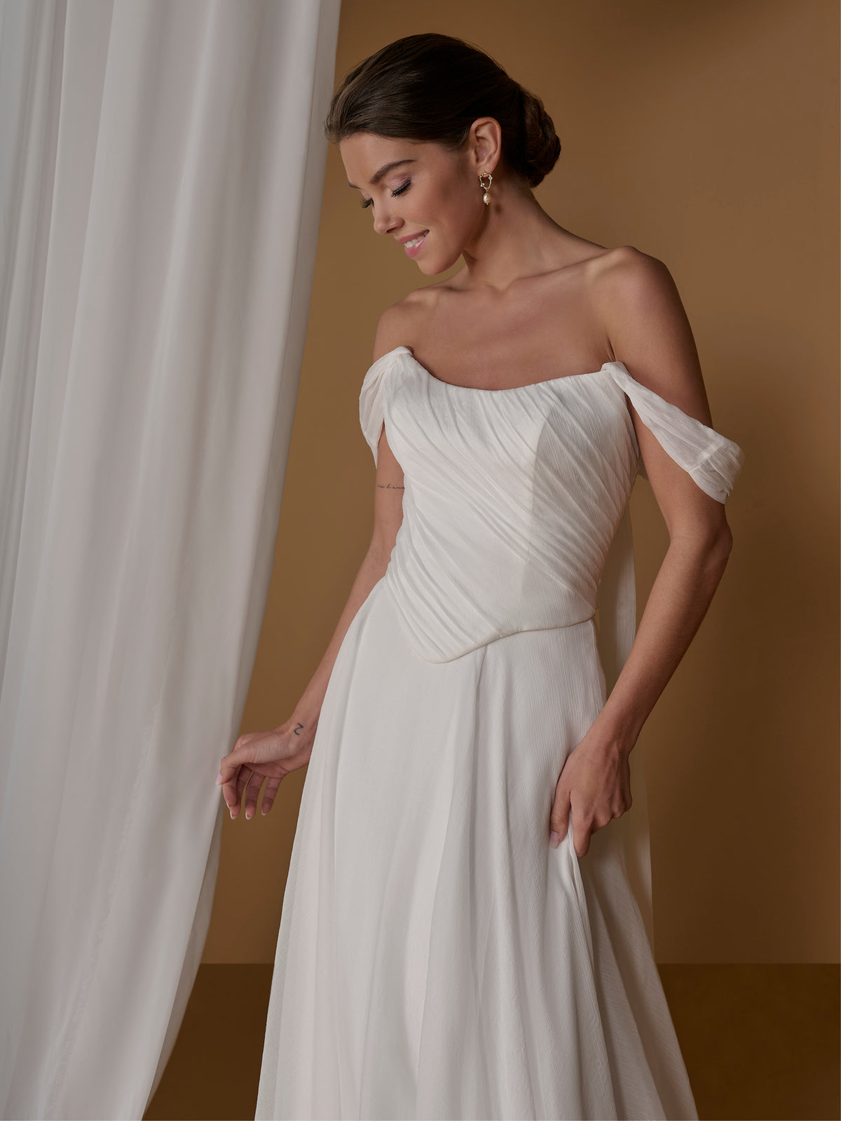 A woman wearing an off-the-shoulder white wedding dress with a gathered bodice and flowing skirt, posed against a warm brown backdrop with a sheer white curtain on the left. / 2-piece crinkle chiffon gown with draped bodice and full skirt.