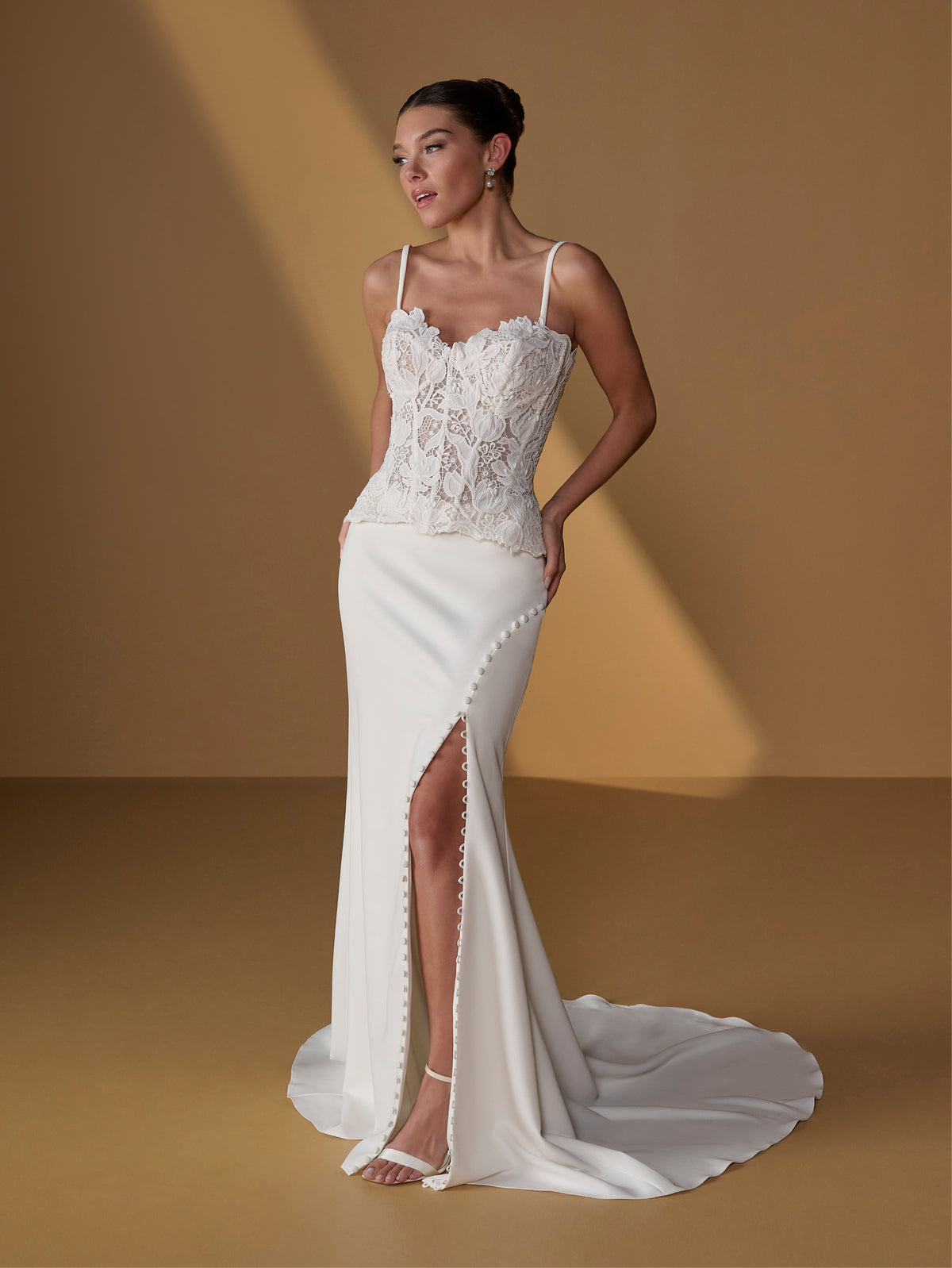 A woman models a white lace and satin wedding gown with thin straps, a fitted lace bodice, a high thigh slit, and a long train against a warm tan studio backdrop. / Lace bodice with lace up back, crepe skirt with side front split.