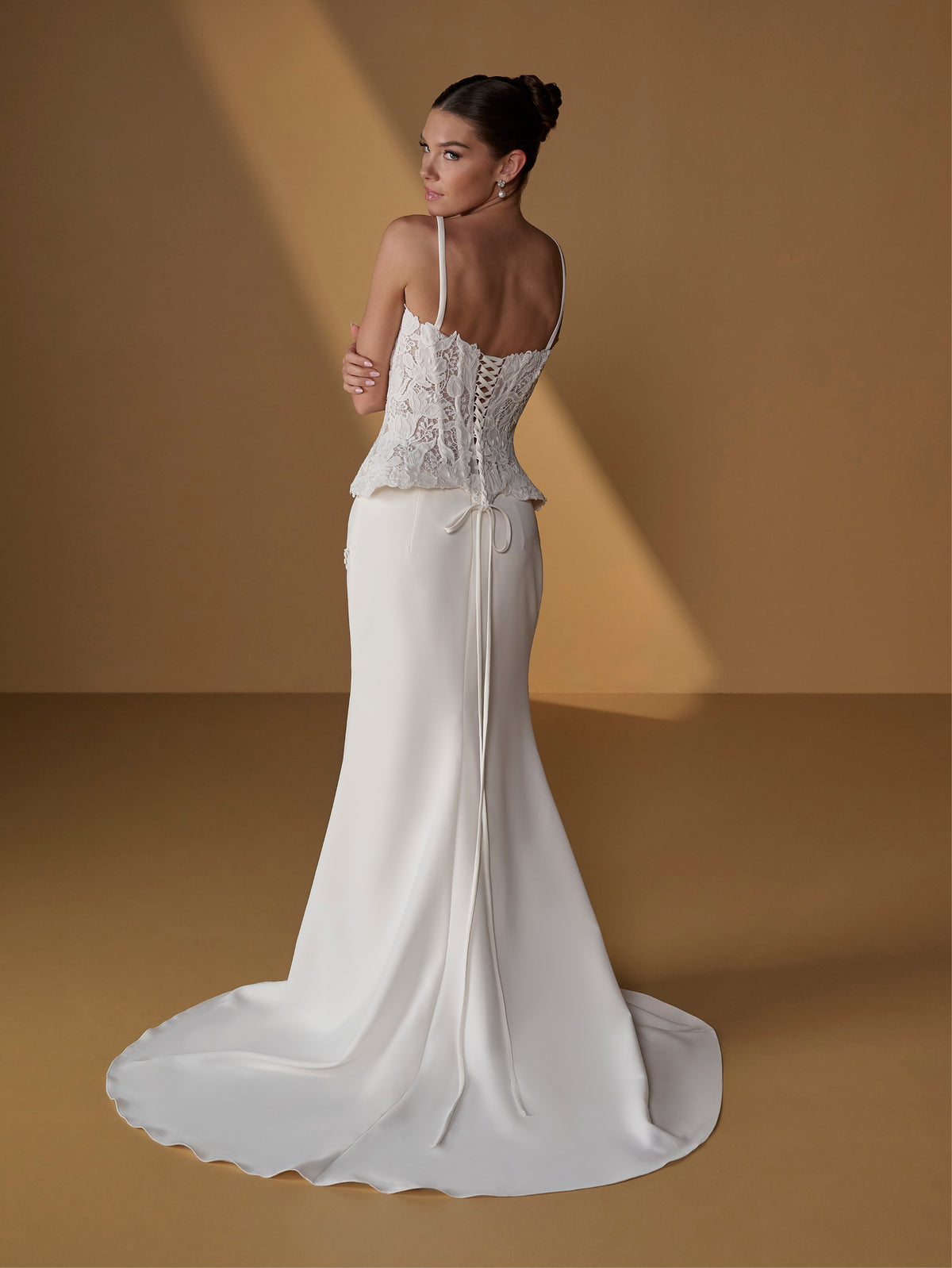 A woman modeling a white bridal gown from the back, standing on a warm tan studio floor with dramatic diagonal lighting. The dress has thin straps, a lace corset-style upper section, a fitted skirt, and a long train. / Lace bodice with lace up back, crepe skirt with side front split.