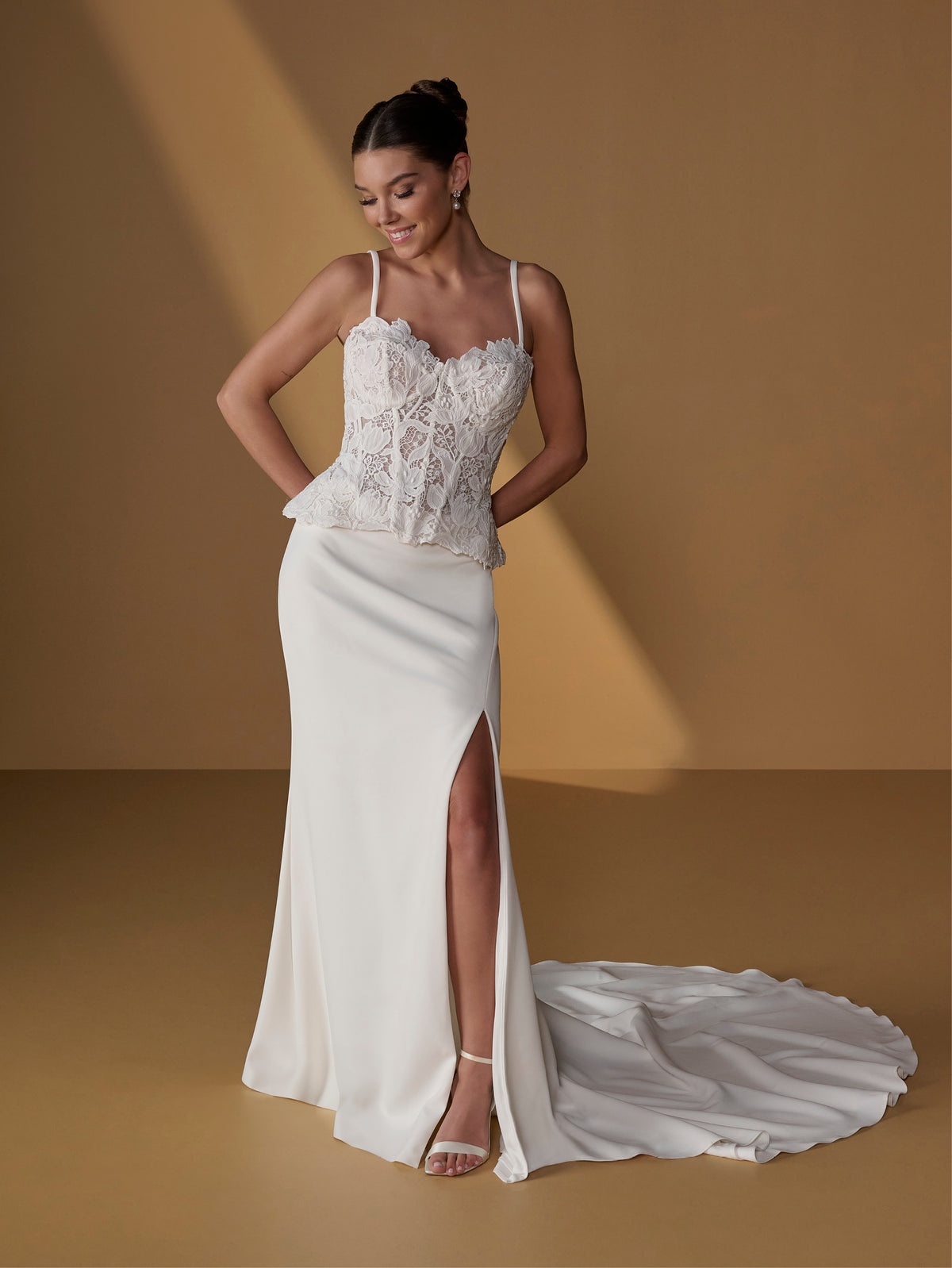 A woman models a white wedding gown with a lace corset-style bodice, thin straps, a fitted satin skirt, and a high front slit. The dress has a long train that pools on the floor against a warm tan studio backdrop. / Lace bodice with lace up back, crepe skirt with side front split.