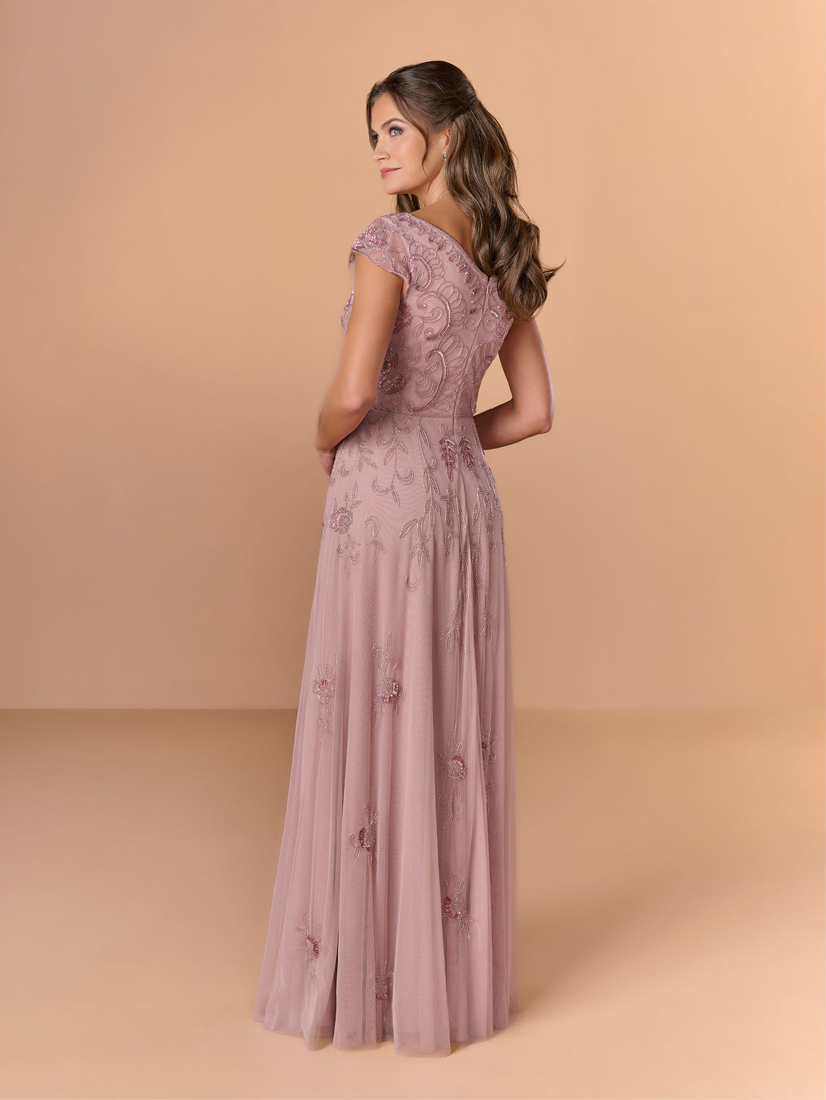 A woman modeling a floor-length dusty pink gown with floral lace embroidery and sheer cap sleeves, shown from the back against a warm beige studio backdrop. / All over beaded gown with sheer cap sleeve, v neckline and full, A-line skirt.