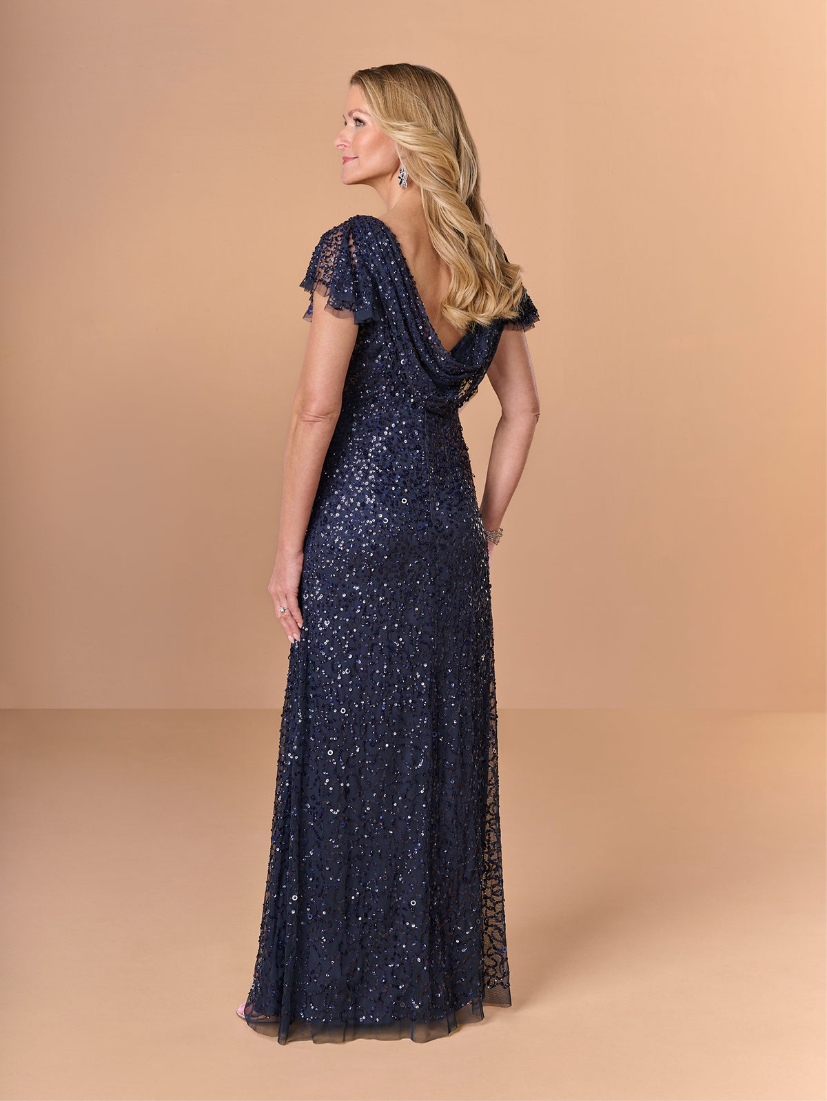 A woman modeling a floor-length navy blue evening gown with sequins and lace, shown from the back against a solid beige studio backdrop. / All over crunchy bead gown with flutter sleeve and cowl back.
