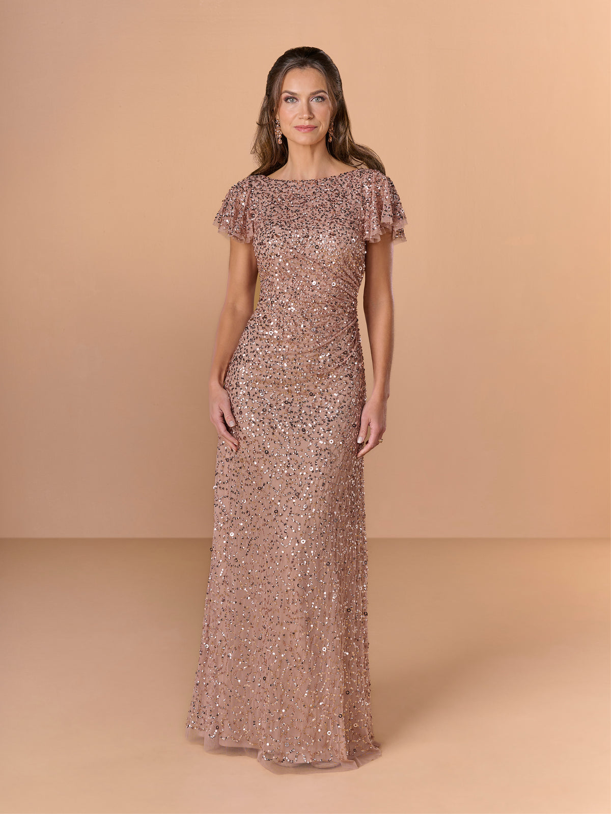 Woman wearing a rose gold sequin gown against a beige background / All over crunchy bead gown with flutter sleeve and cowl back.