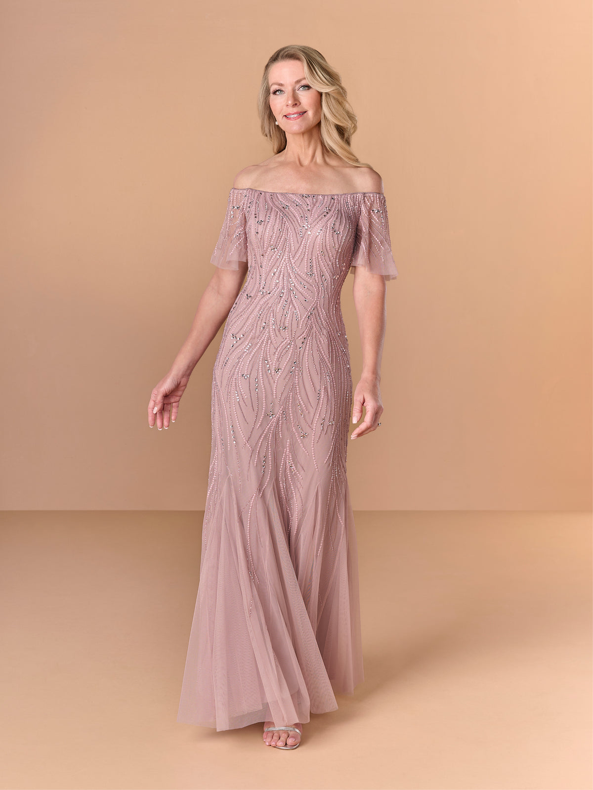 A woman modeling an off-the-shoulder floor-length gown in dusty blush pink with intricate bead and sequin embellishments, set against a solid warm beige studio background. / All over beaded gown with chic off the shoulder neckline and soft godet skirt.