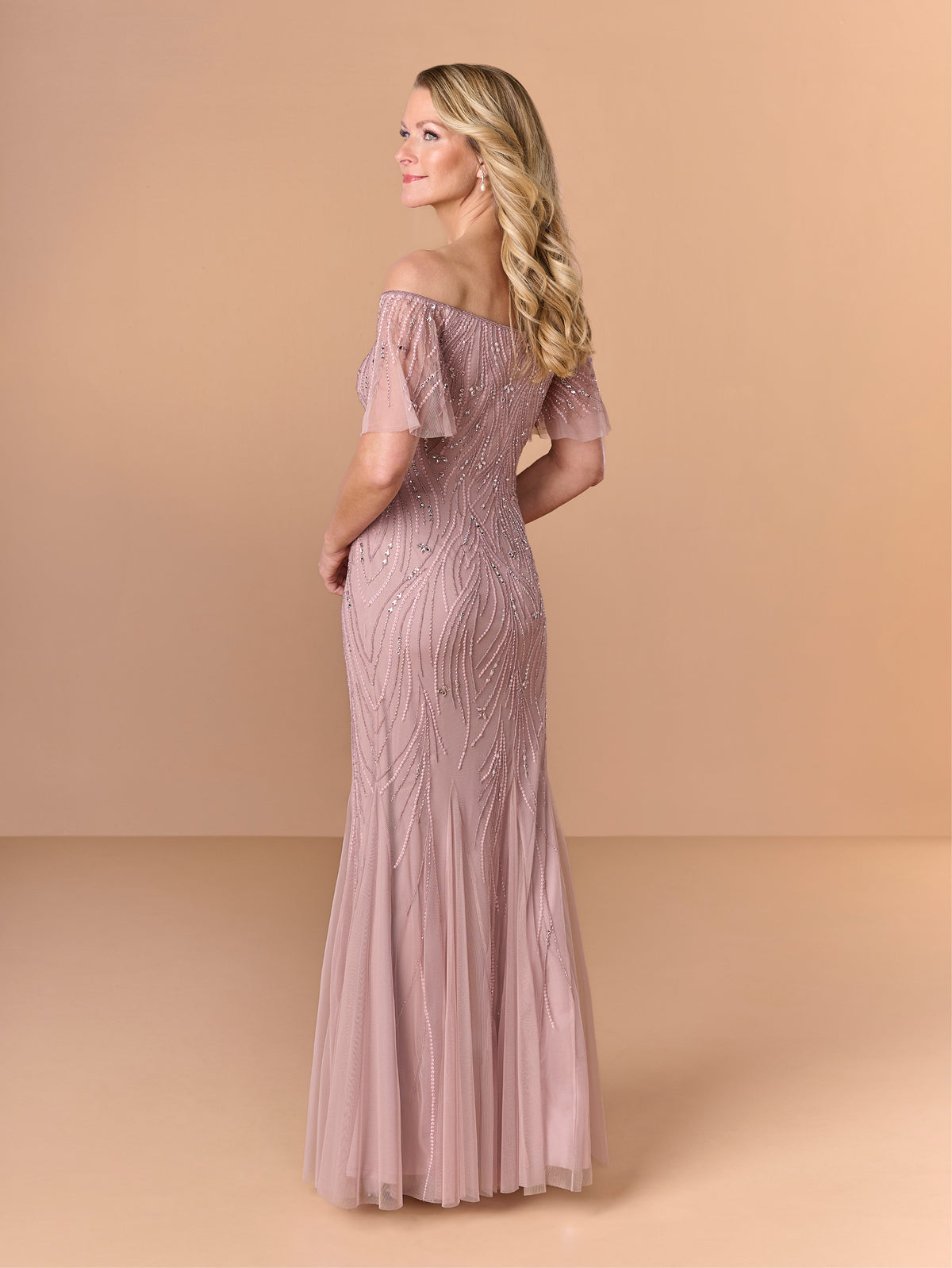 A woman modeling an off-the-shoulder floor-length gown in a dusty rose pink color, featuring intricate beadwork and tulle detailing, set against a solid beige studio background. / All over beaded gown with chic off the shoulder neckline and soft godet skirt.