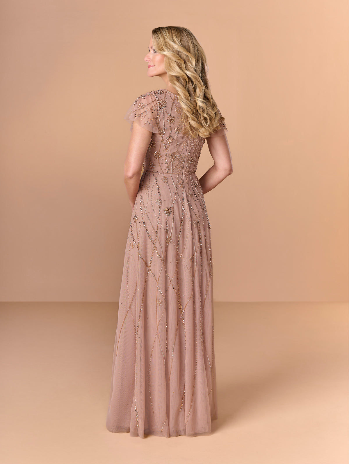 A woman wearing a floor-length dusty rose formal gown with intricate beadwork and sequin embellishments, shown from the back against a solid beige studio backdrop. / All over beaded gown with v neckline and soft A-line skirt.