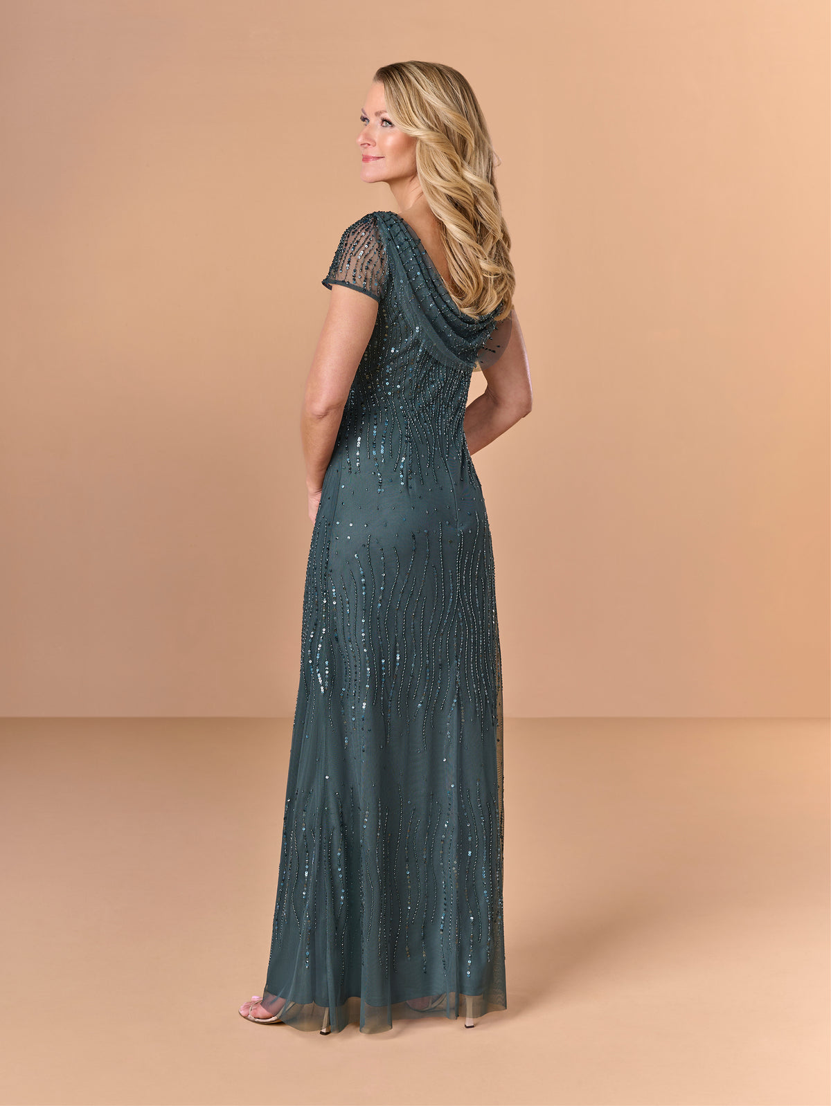 Woman wearing a long, dark green dress with sequin details on a beige background / All over beaded gown with body shirring, skirt slit and back cowl detail.