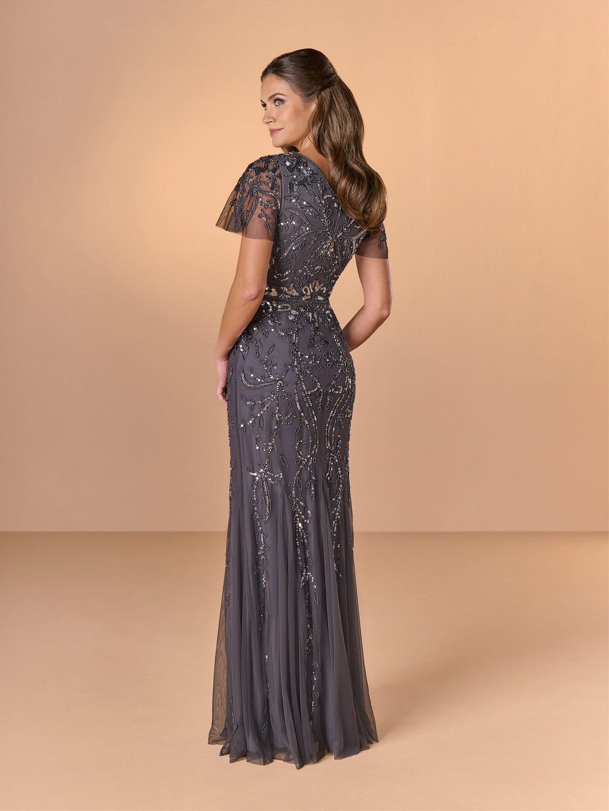A woman modeling a floor-length evening gown from the back, featuring sheer short sleeves, a fitted waist, and intricate beaded embellishments on a dark gray fabric, set against a solid beige studio backdrop. / All over beaded gown with short flutter sleeve and soft godet skirt.