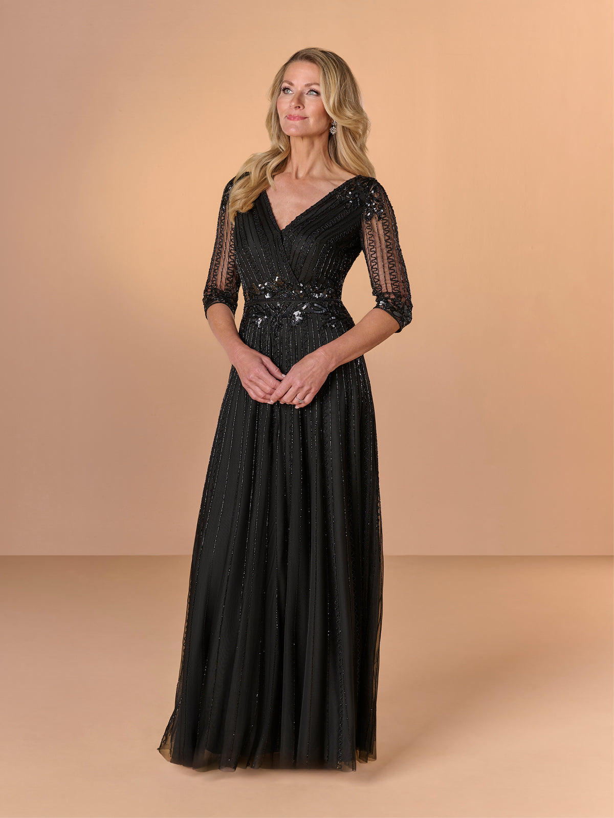 Woman wearing a black evening gown with sheer sleeves against a beige background / All over beaded gown with surplice bodice, elbow length sleeve and full A-line skirt.