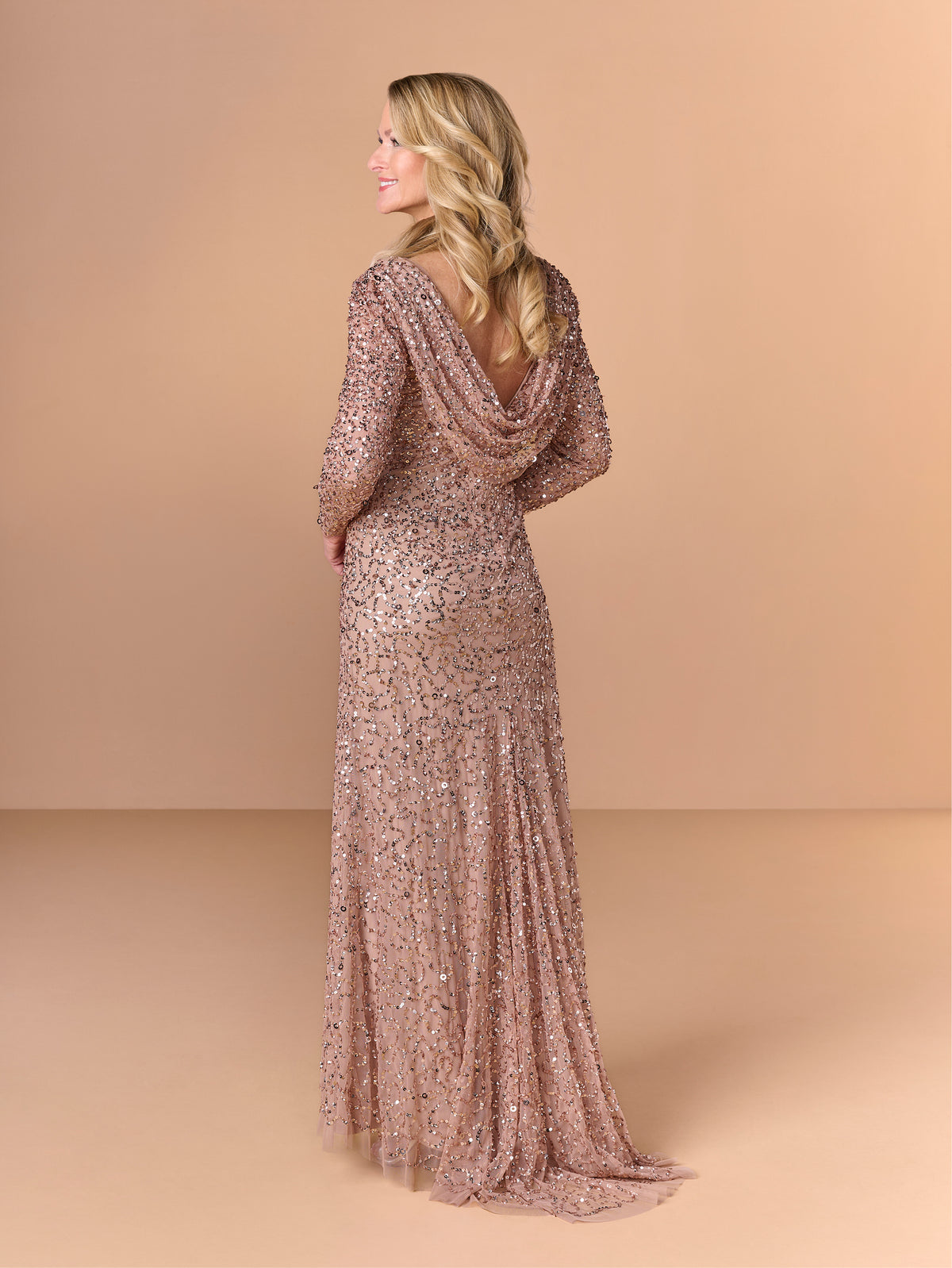 A woman modeling a floor-length, long-sleeve evening gown with a draped cowl neckline and dense sequin embellishment, shown from the back against a warm beige studio backdrop. / All over crunchy bead gown with long sleeves and cowl back.